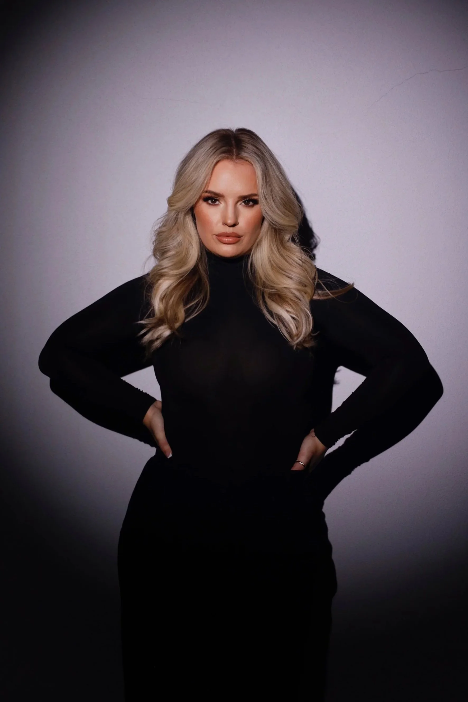 Blonde woman with styled hair wearing a black long-sleeve top and black pants, standing with hands on hips against a gradient background.  Natasha Wilkes, owner and founder of the Aesthetic Ranch in Fort Lauderdale.