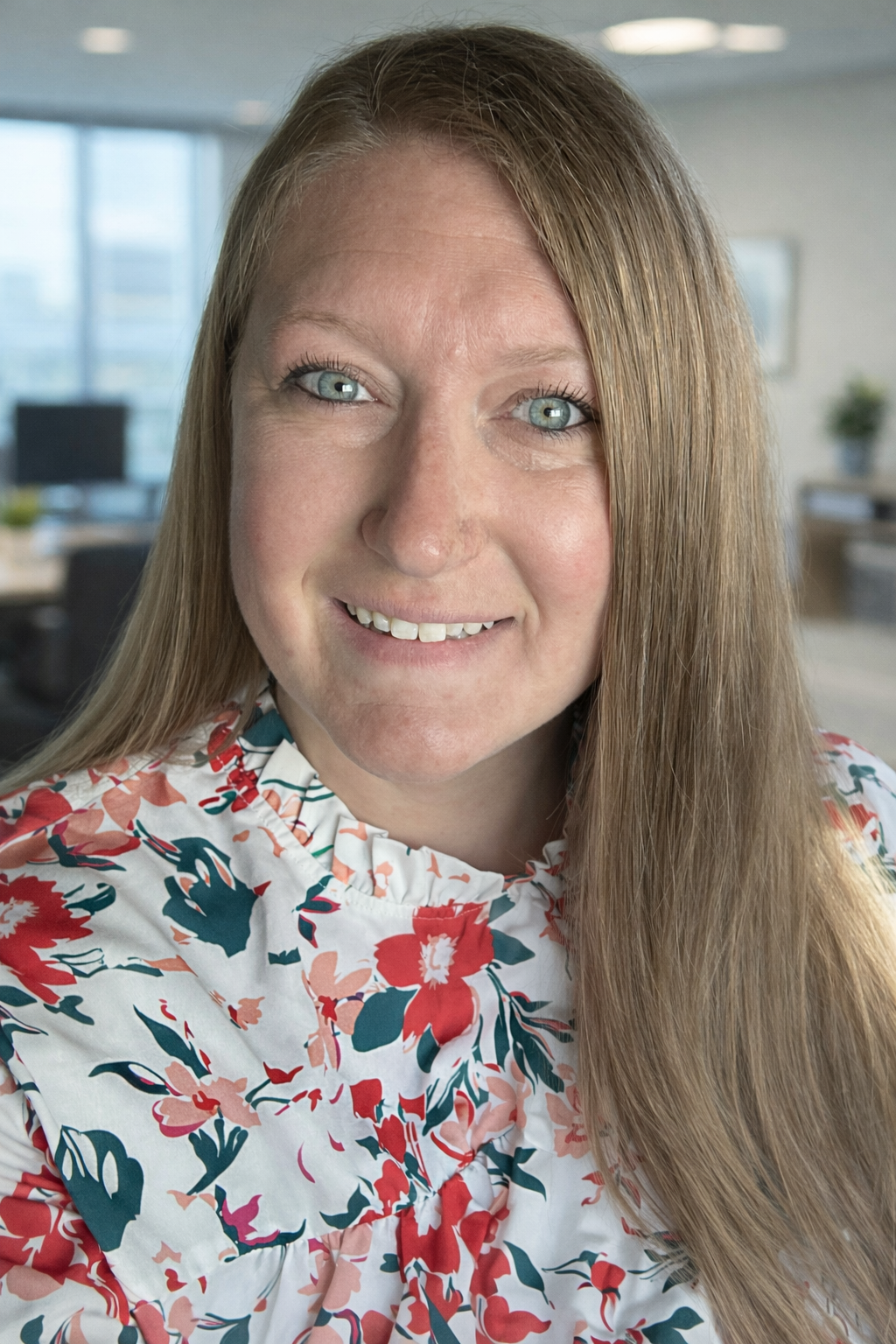 Close-up portrait of a woman with long red hair and blue eyes, smiling in an office setting with large windows and modern decor.