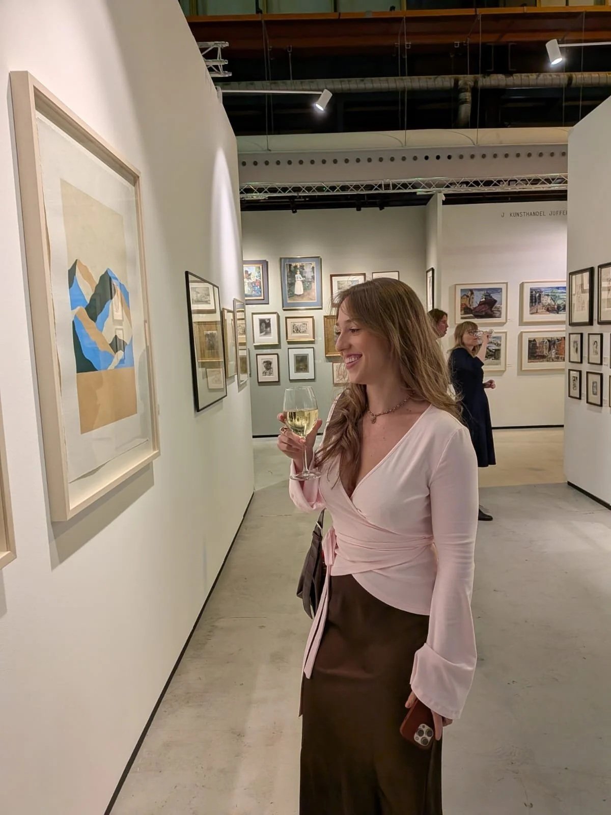 A woman with long brown hair, wearing a light pink wrap blouse and dark brown skirt, is smiling and holding a glass of white wine at an art gallery with white walls and artworks displayed.