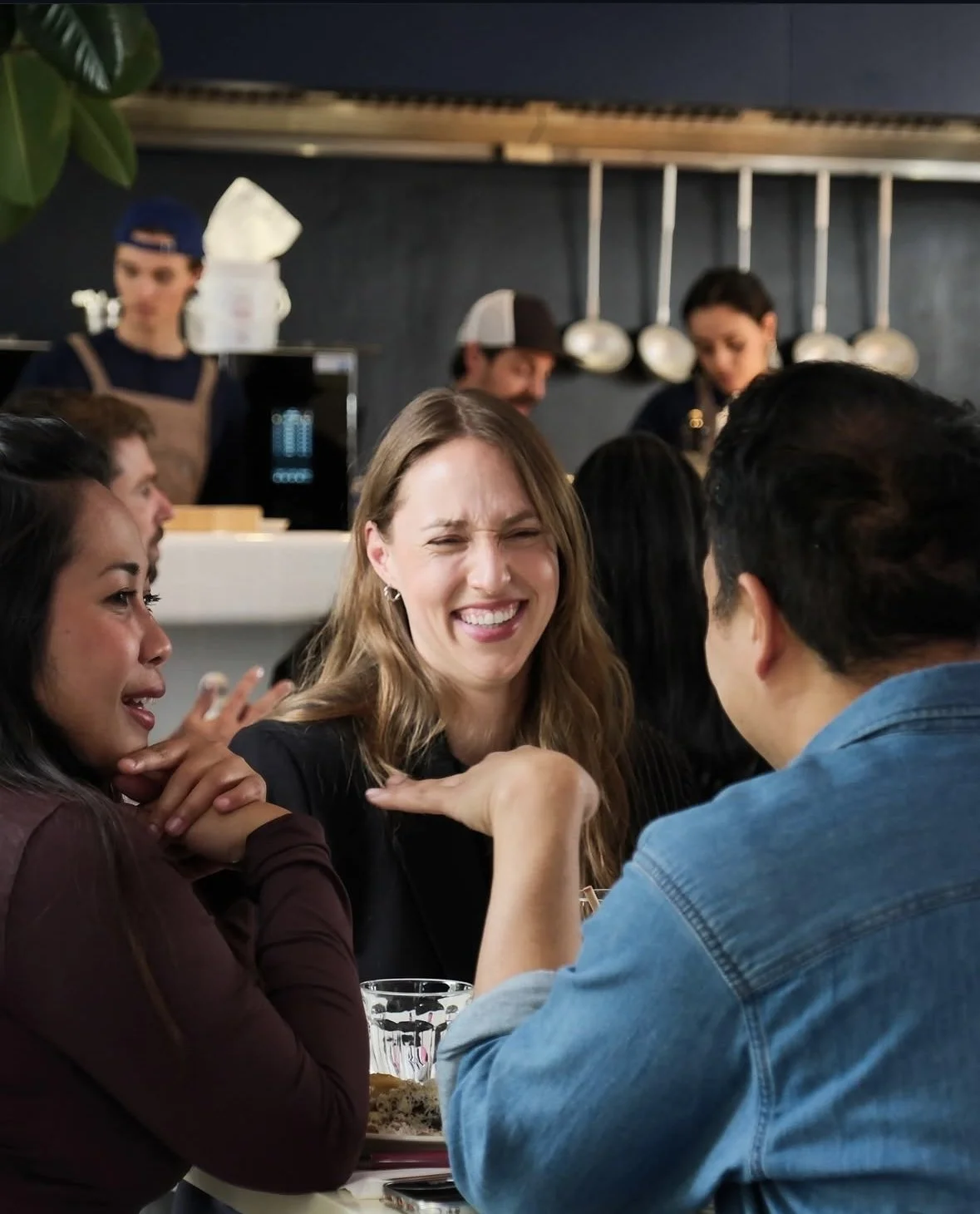 A group of people enjoying a lively conversation at a restaurant, with a woman smiling and two others engaged with her. In the background, restaurant staff and hanging kitchen utensils are visible.