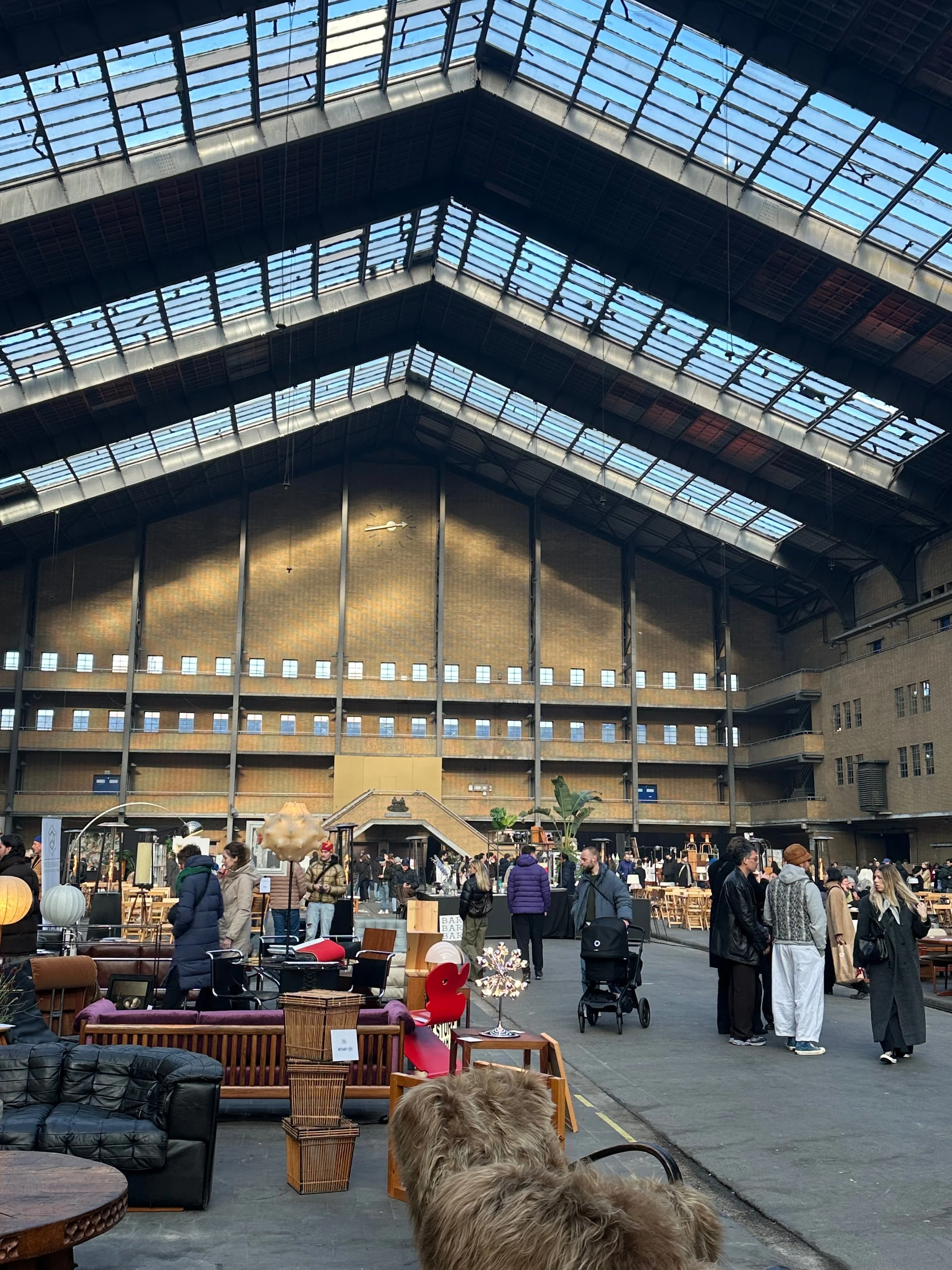 Indoor market or event space with high glass ceiling, people walking and browsing furniture and decor items, some people with strollers, and various furniture and decorative objects on display.
