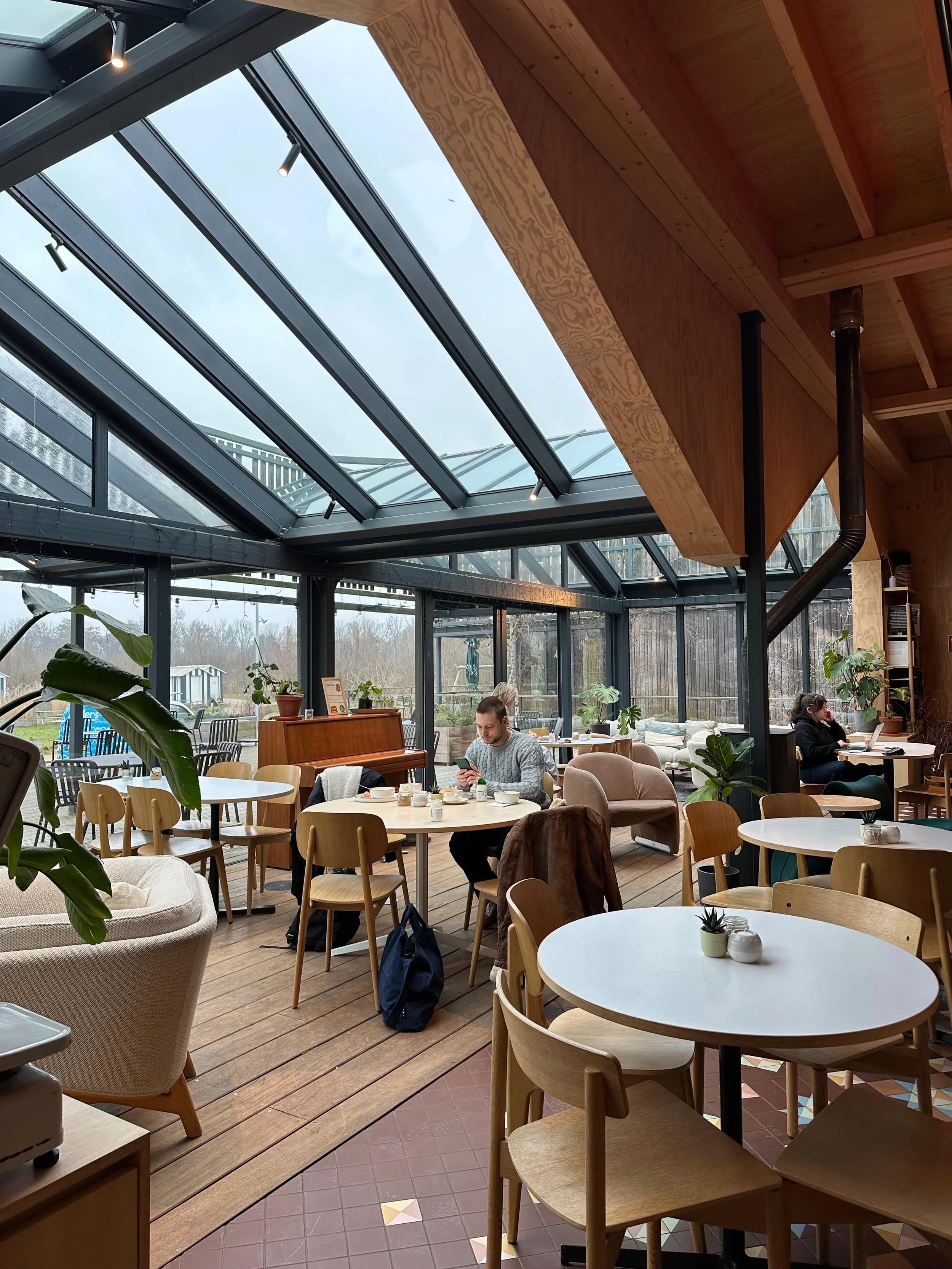 Cafe interior with wooden ceiling, large glass skylight, and patrons sitting at tables, some using electronic devices.
