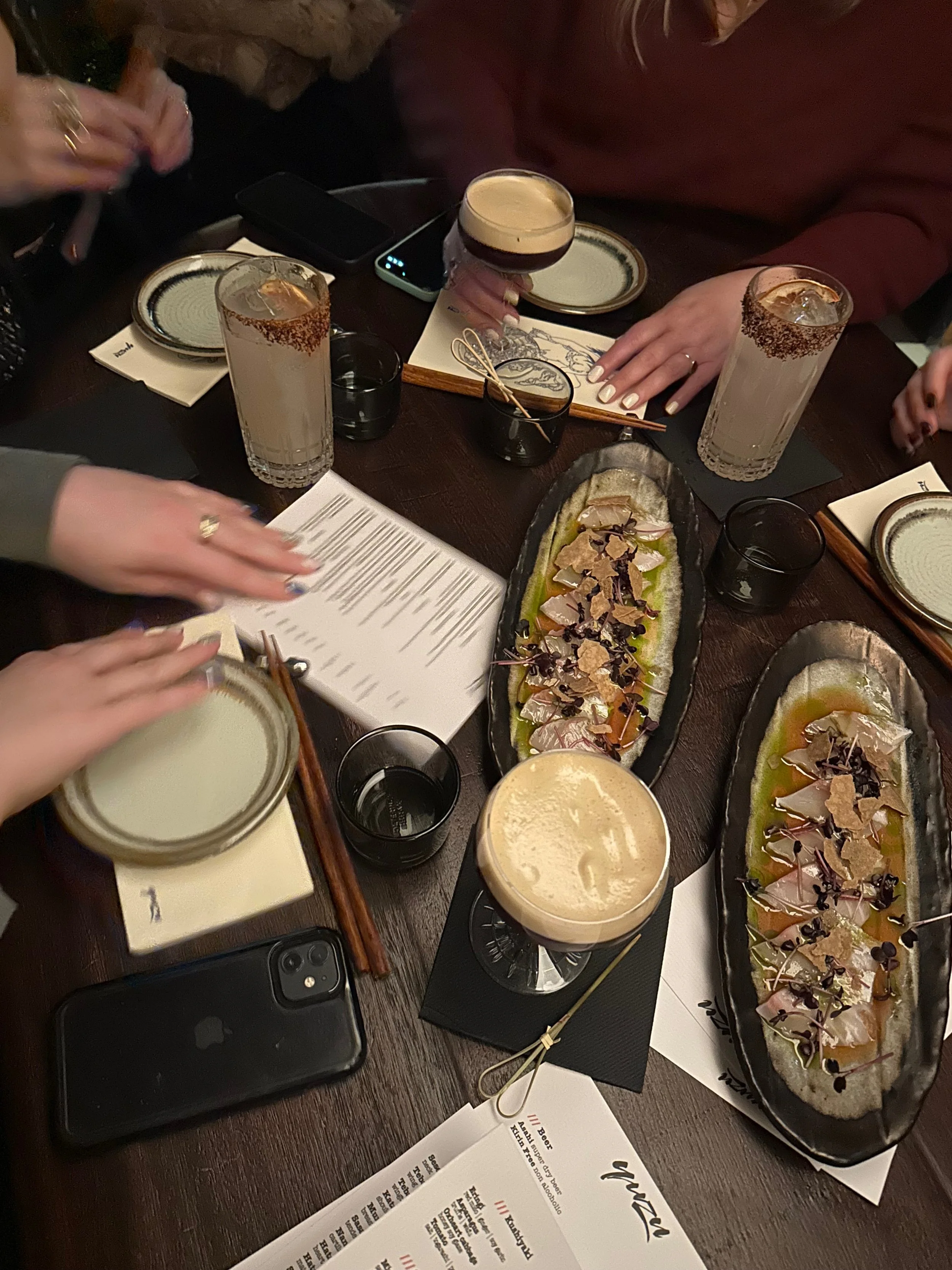A group of people sitting around a dark wooden table with plates of sashimi, glasses of cocktails, and a menu, during a dinner gathering.