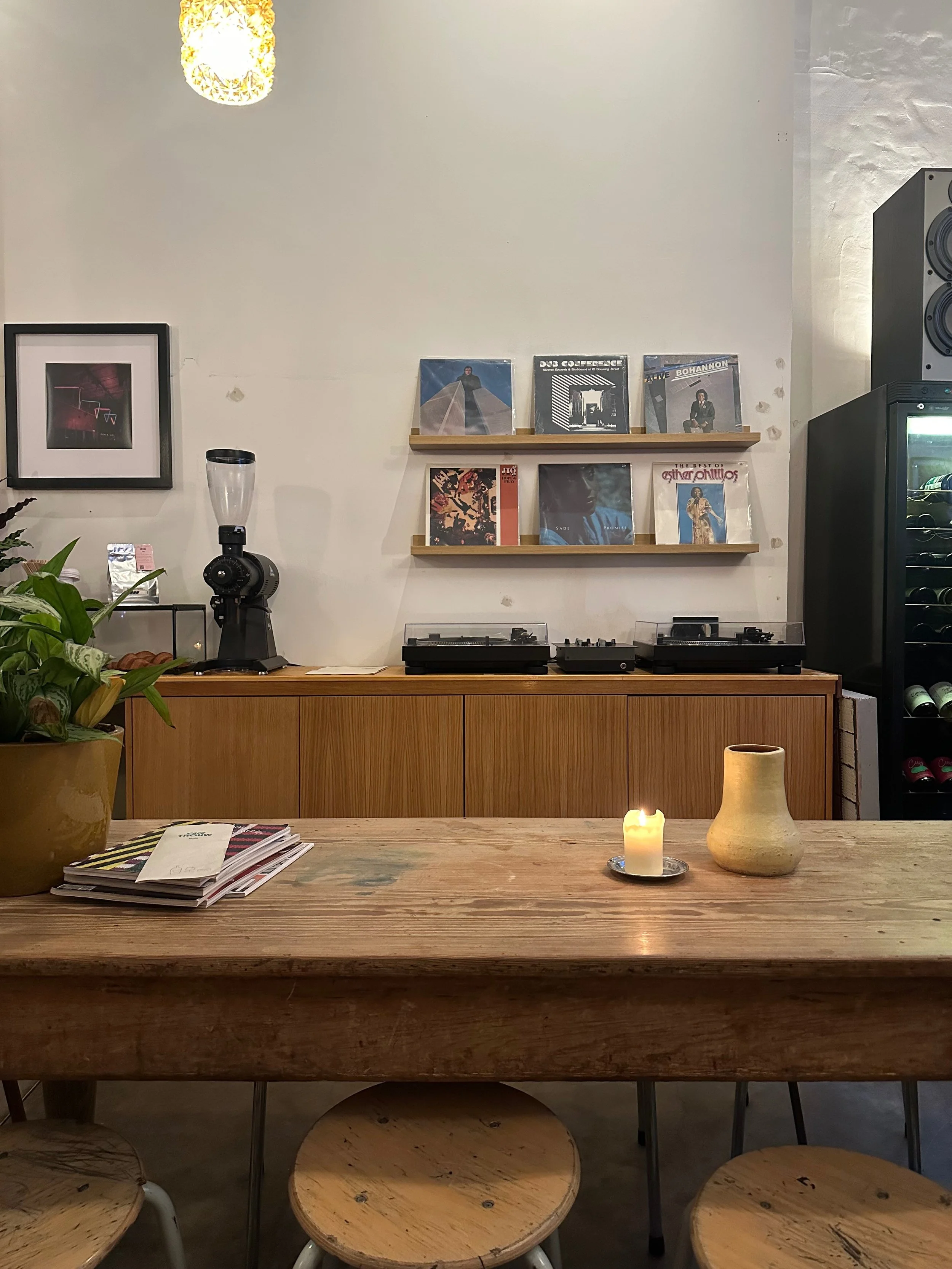 A cozy interior space with a wooden dining table, candles, a potted plant, and a wall-mounted vinyl record display, featuring turntables, vinyl records, and a wine cooler.