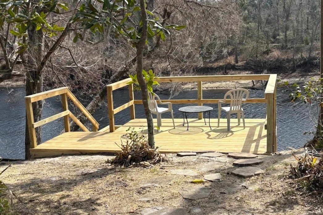 A wooden deck with a railing, two plastic chairs, and a small round table, situated by a river with trees in the background.