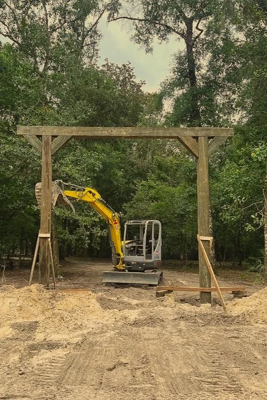 A small yellow excavator working on construction, positioned in front of a wooden frame structure with dirt ground in the foreground and a forest background.