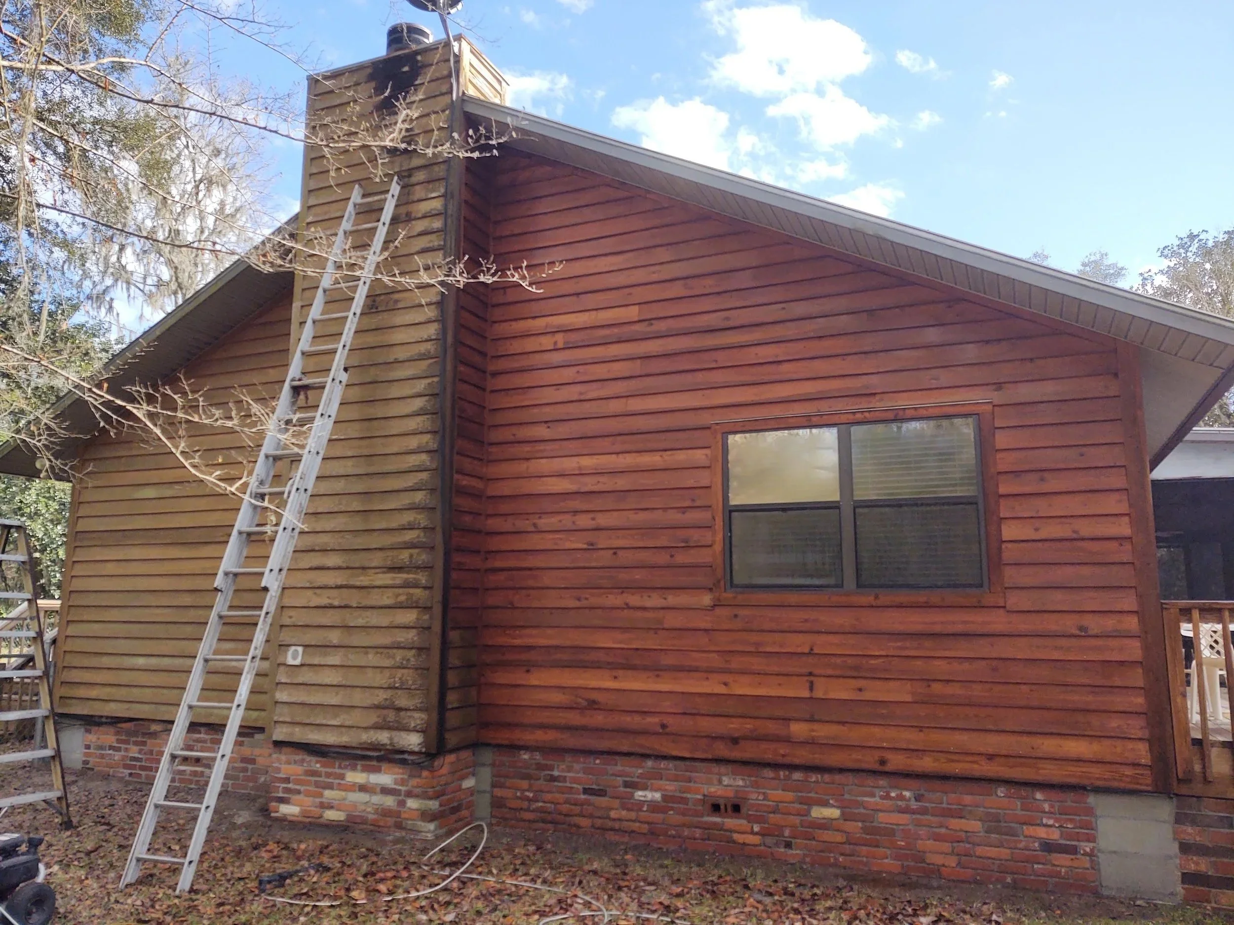The back of a house with a section being renovated or cleaned, featuring wooden siding, a ladder, and a chimney. The house has a brick foundation, a window, and a sloped roof. There are trees and a blue sky in the background.
