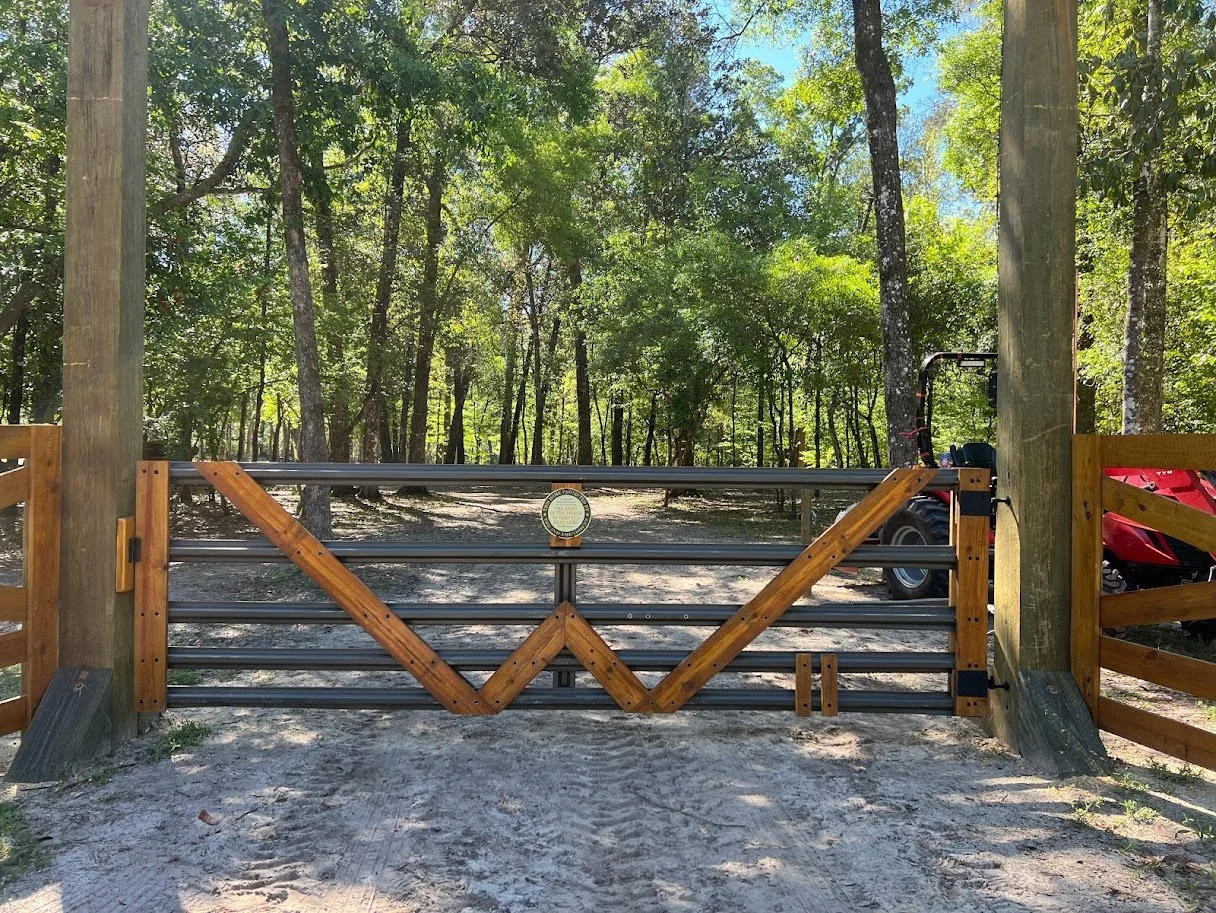 A wooden gate with metal bars blocking a dirt road in a wooded area, with trees and sunlight overhead.
