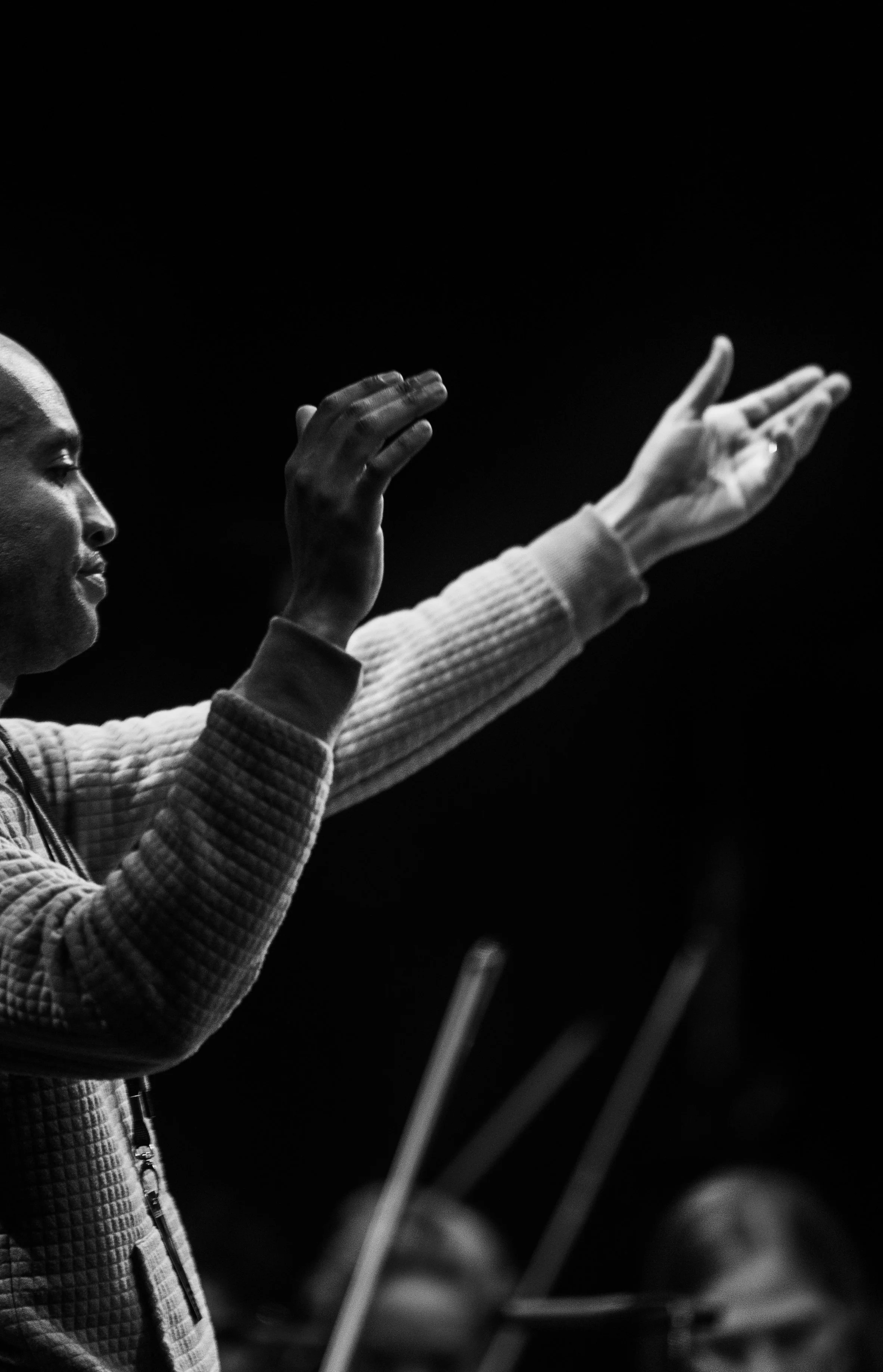 Black and white image of a man with dark skin raising his right hand with an open palm while looking at something off-camera. He's wearing a textured, quilted jacket and has a focused expression.