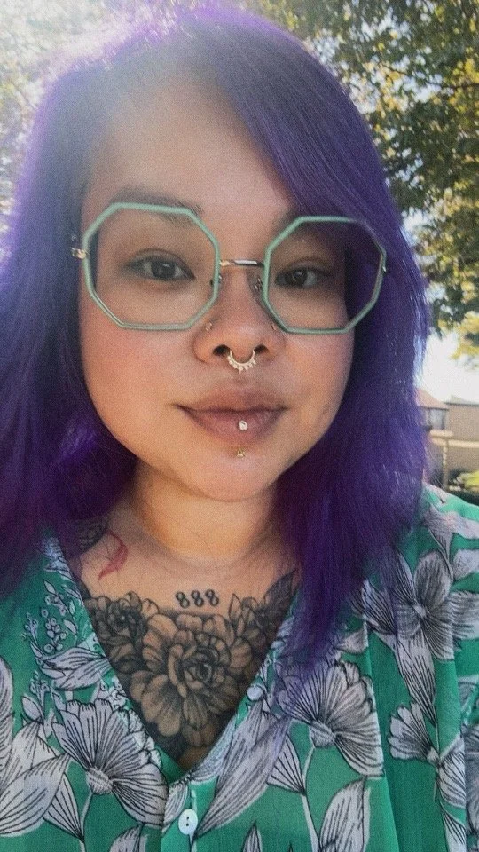 Young woman with purple hair, wearing geometric glasses, and multiple facial piercings, standing outdoors with trees in the background.