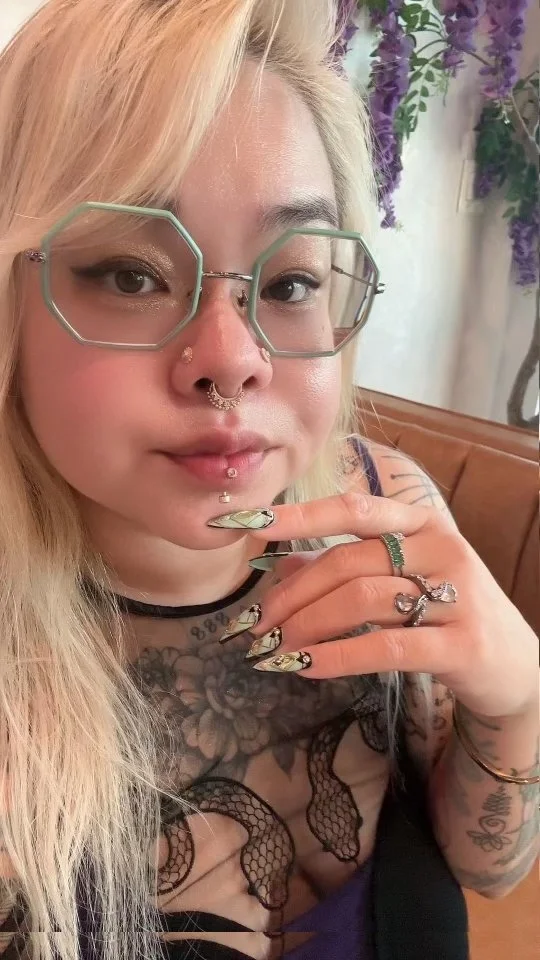 with platinum blonde hair wearing large, green octagonal glasses, a nose ring, and multiple lip piercings. She has tattoos on her chest, arms, and fingers, and is wearing rings and long, decorated nails. sitting on a brown leather bench with a backdrop of purple flowers.
