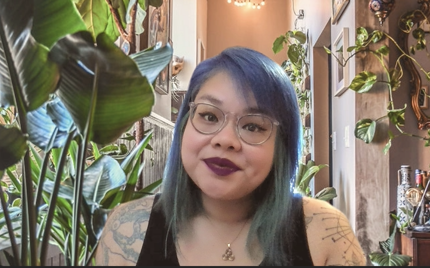 Jax NTP with blue and green hair, wearing glasses, dark lipstick, and a black top, surrounded by green plants in a cozy, decorated indoor space.