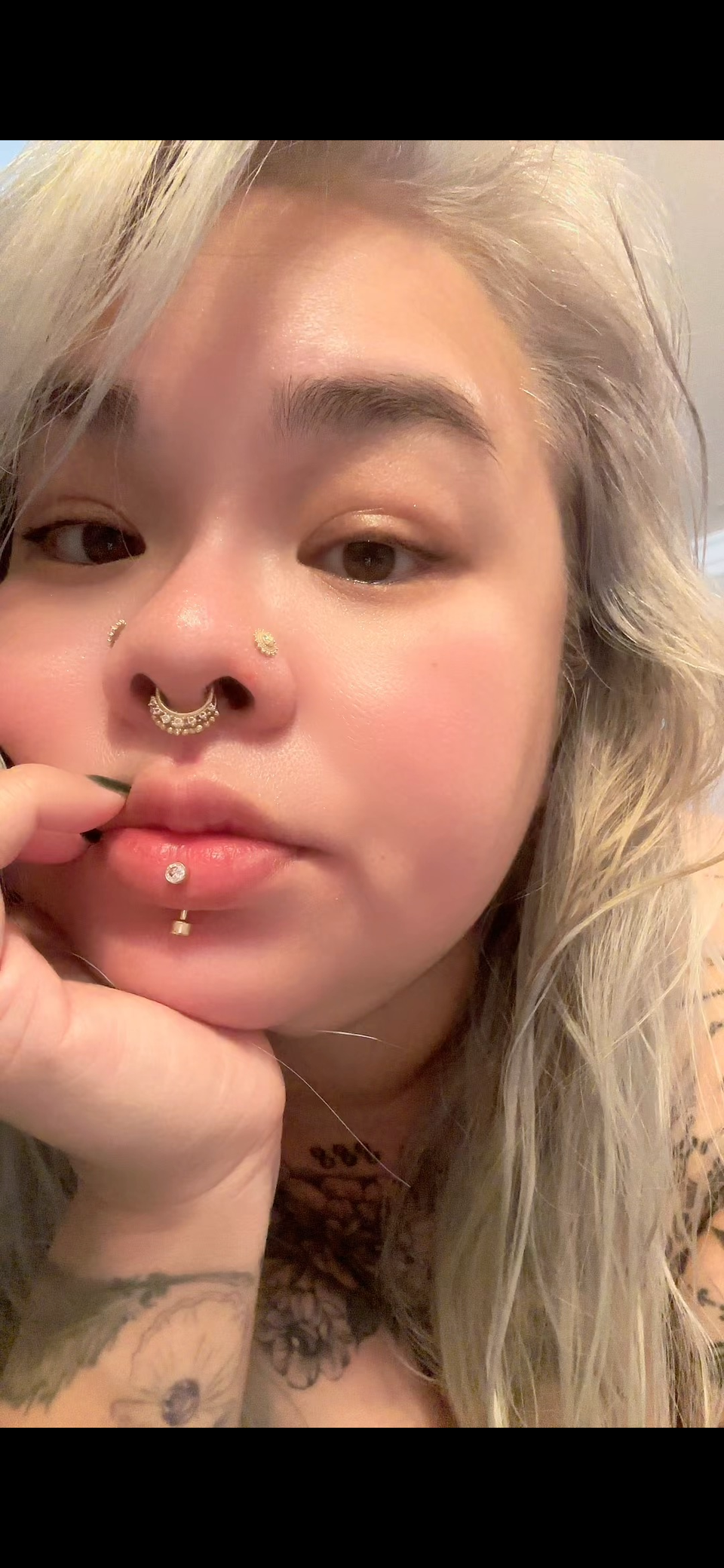 Jax NTP with blonde hair and multiple facial piercings, including a septum ring, nostril piercings, and a labret piercing. They have tattoos on their arm and neck, and are holding their hand near their face.