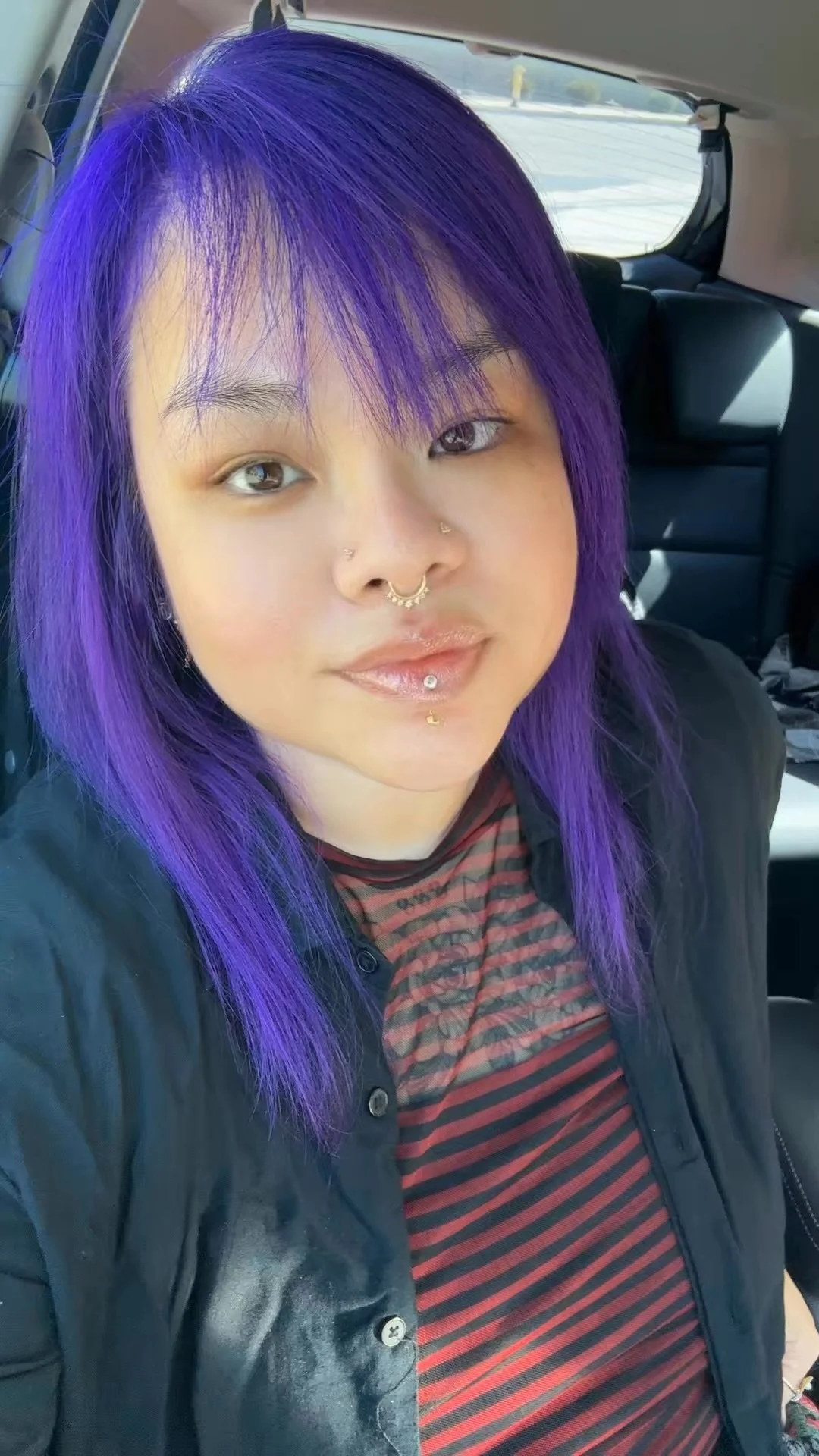 Jax with bright purple hair, multiple facial piercings, and wearing a black button-up shirt over a red and black striped top, sitting inside a vehicle.