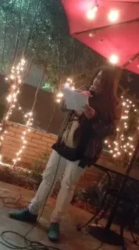 Jax NTP reads - at MFA Reading Series at Wine Crush in Long Beach - into a microphone outdoors at night, under a red umbrella with string lights overhead.