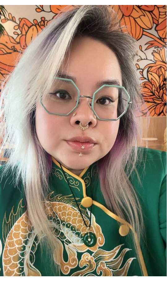 A Jax with blonde, purple-tinted hair wearing hexagonal glasses, a green and gold embroidered jacket, and a necklace with a circular pendant. She has facial piercings, including a septum and lip ring, and a floral patterned wall behind her.