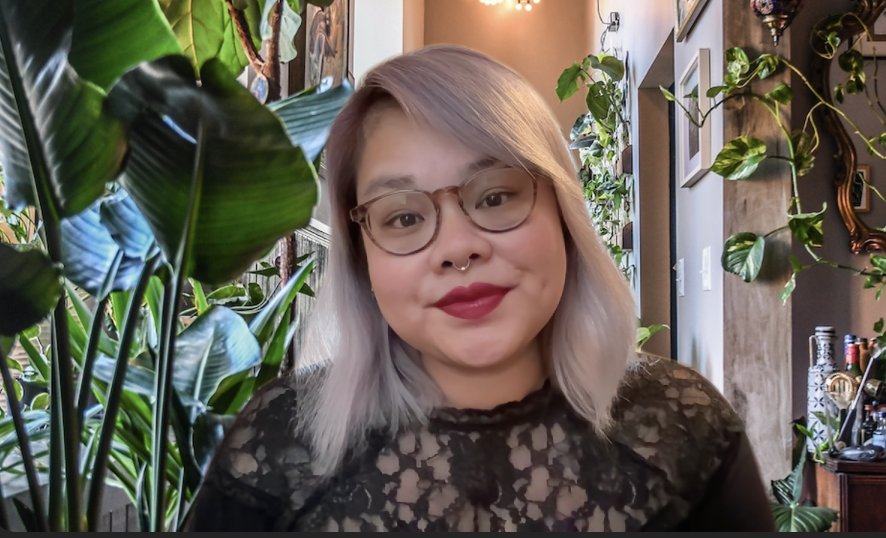 Jax NTP with light blonde hair, glasses, and lipstick smiling in a room filled with green potted plants and home decor.