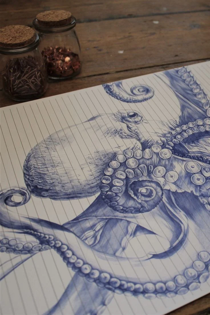 A detailed blue ink drawing of an octopus on lined paper, with jars of colored pencils nearby on a wooden surface.