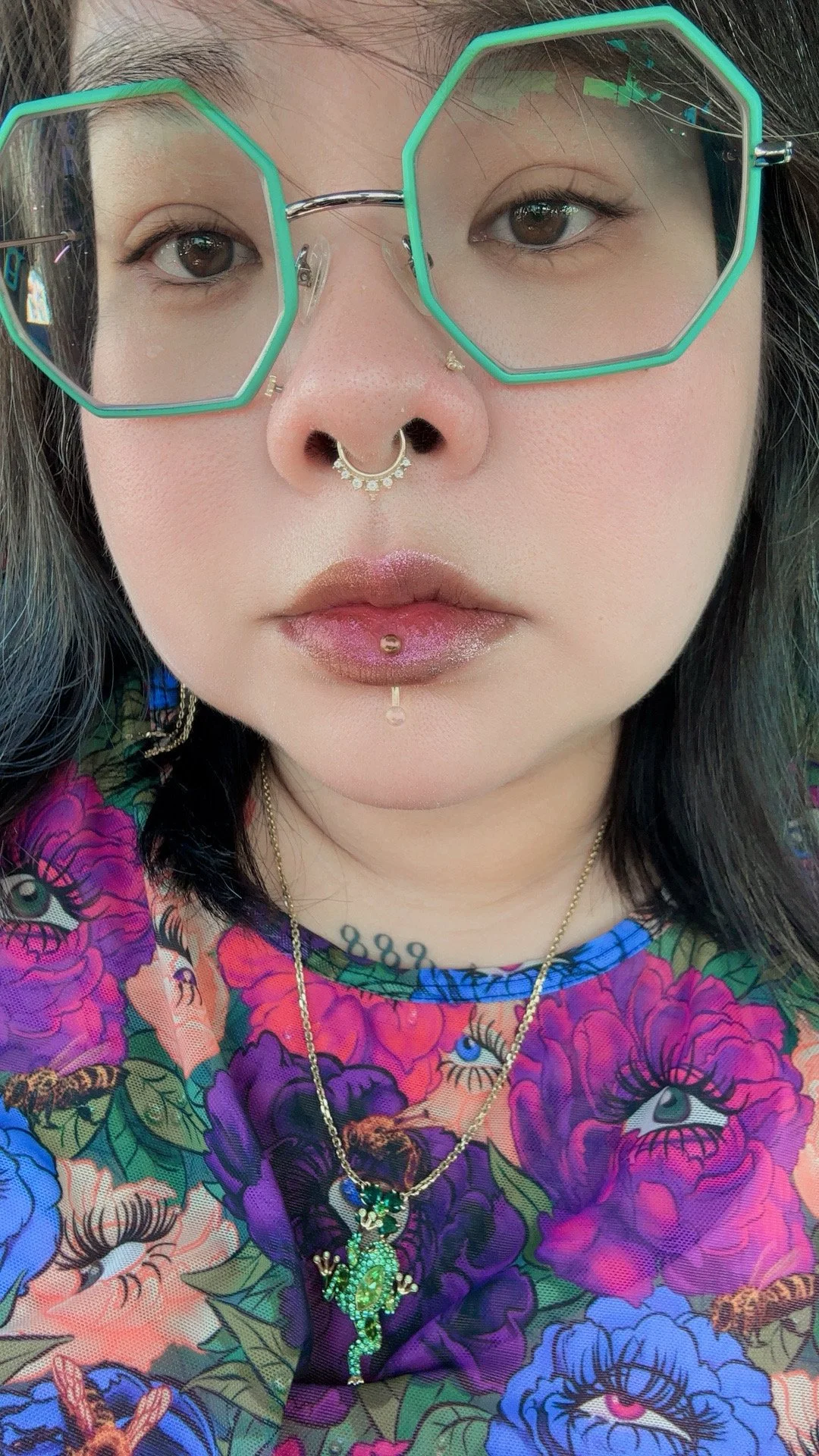 Close-up of a Jax wearing large hexagonal green sunglasses, multiple nose and lip piercings, and a colorful floral shirt with eye and bee patterns. She has dark hair, and jewelry including a necklace with a green frog pendant.