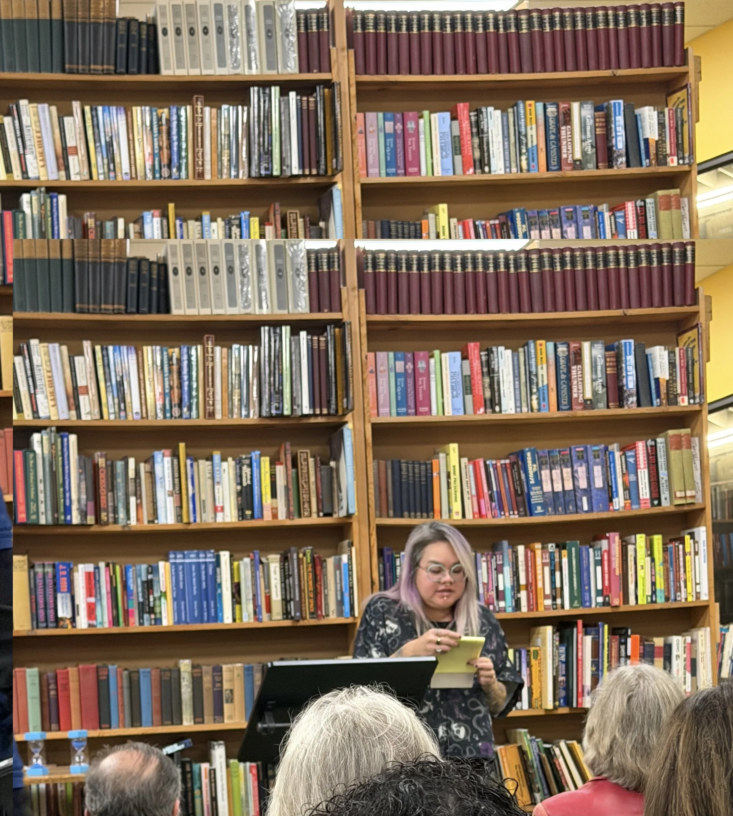 A woman with glasses and dyed purple hair reading from a yellow paper in front of a large bookshelf filled with colorful books, with a group of people listening in a library or bookstore.