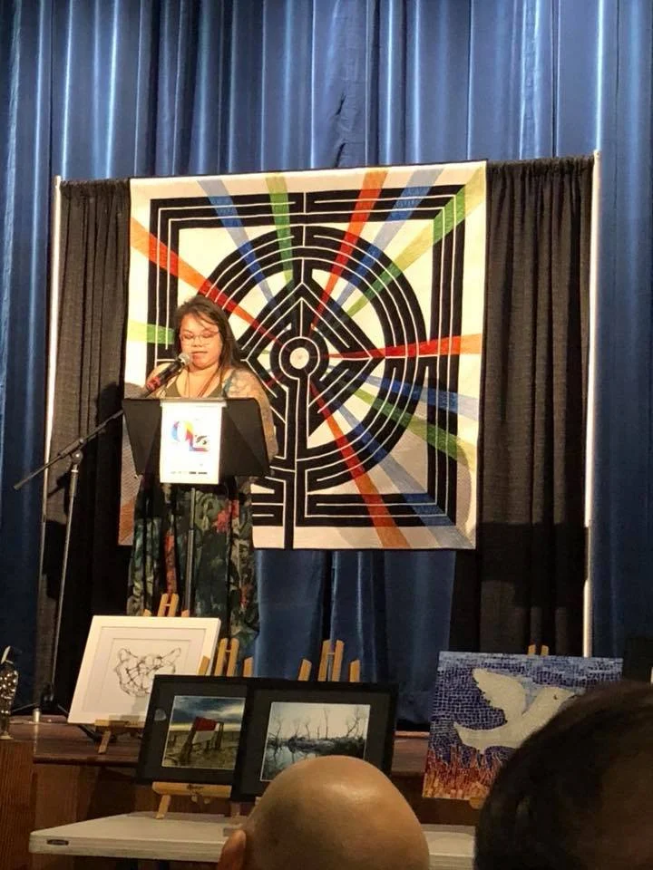 Woman standing at a podium giving a speech or presentation in front of a colorful geometric backdrop. Several framed artworks are displayed on easels in the foreground.