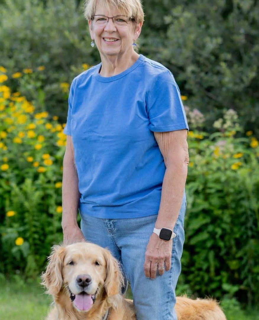 An elderly woman smiling outdoors in a garden with yellow flowers, wearing a blue t-shirt, jeans, glasses, and a smartwatch, standing next to a golden retriever with its mouth open.