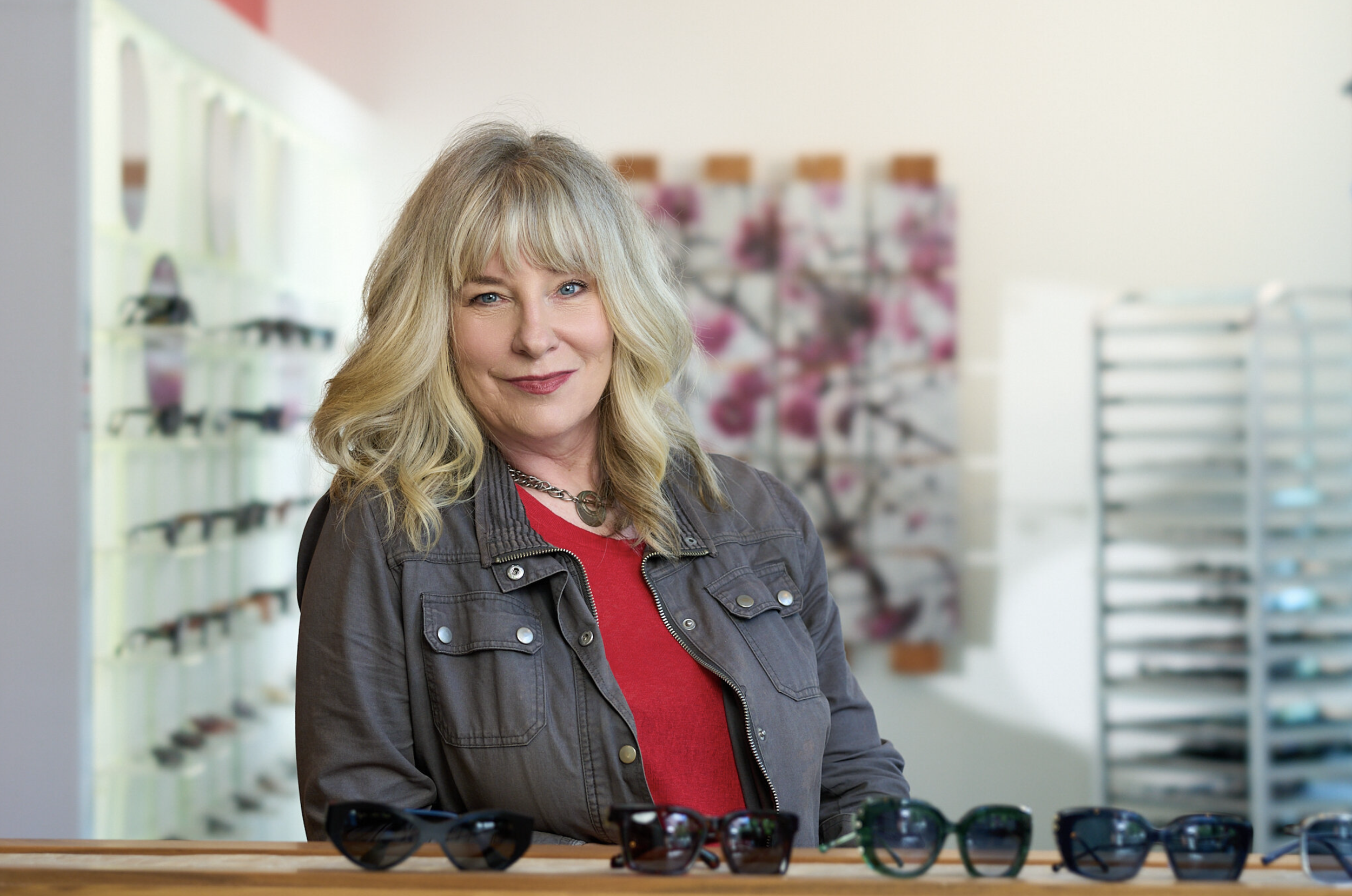 The owner of Visage Eyewear, Tennys Enders, wearing a black jacket over a red shirt, standing behind a table with several pairs of sunglasses in front of her. Background shows a retail store interior with blurred shelves and displays.
