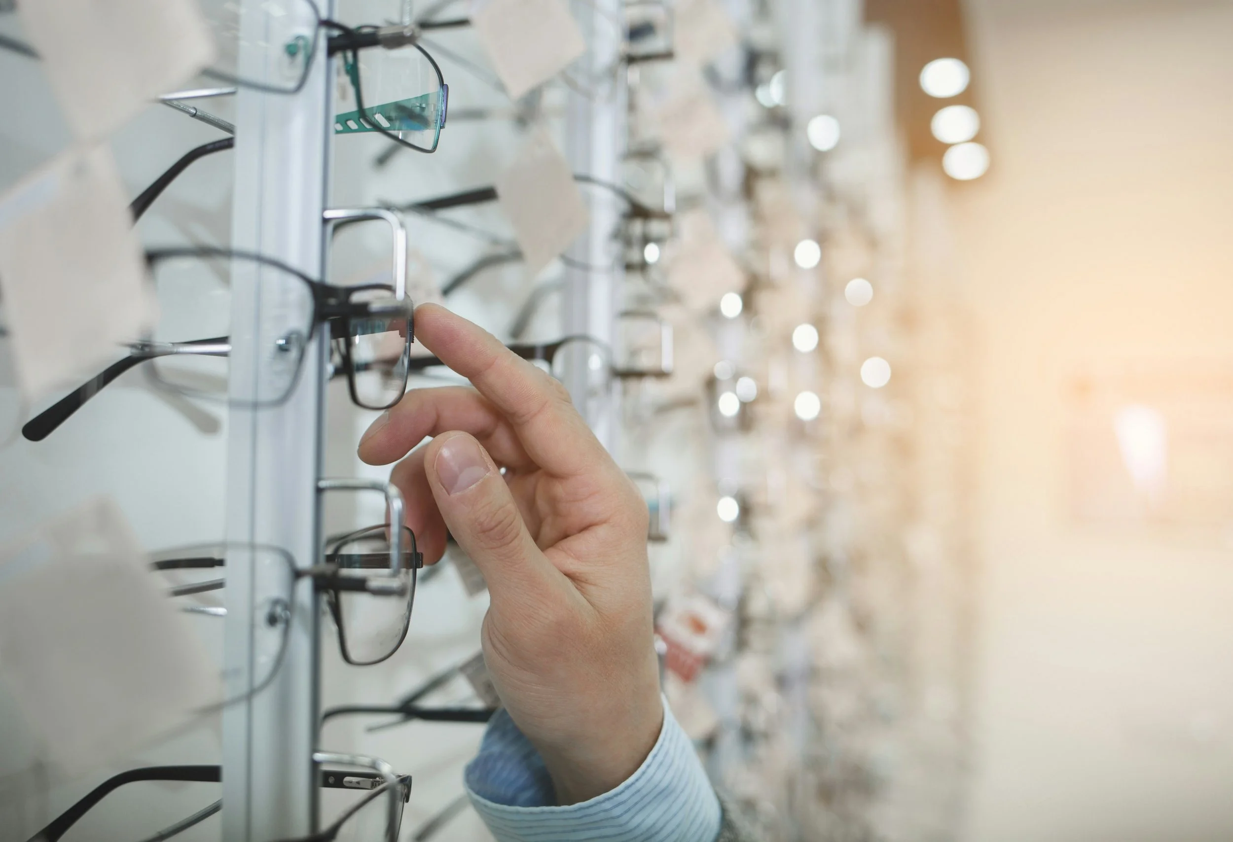 Person browsing eyeglasses in an optical store, holding a pair of black-framed glasses with their right hand.