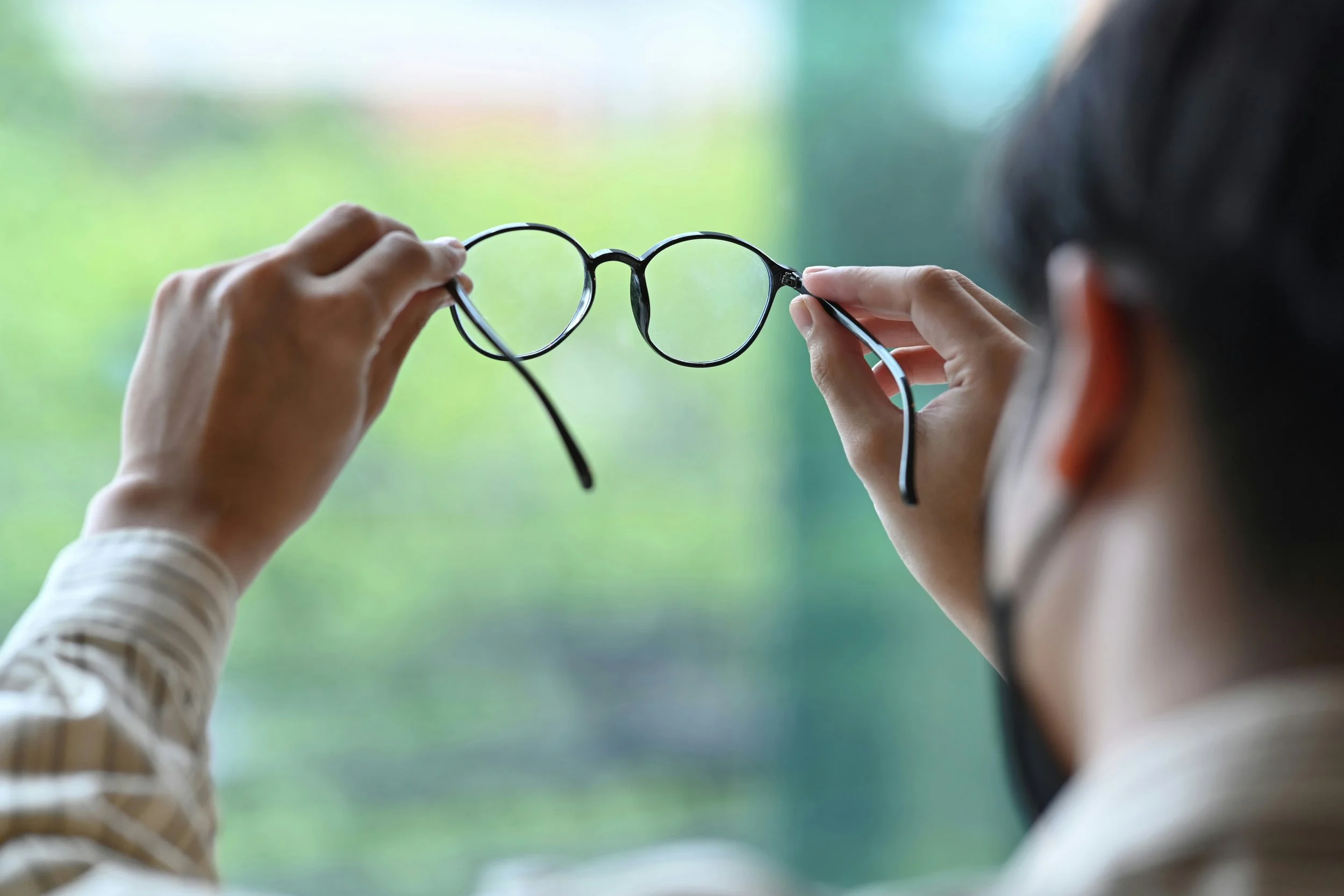 Person holding a pair of black round glasses in front of a blurry green background.