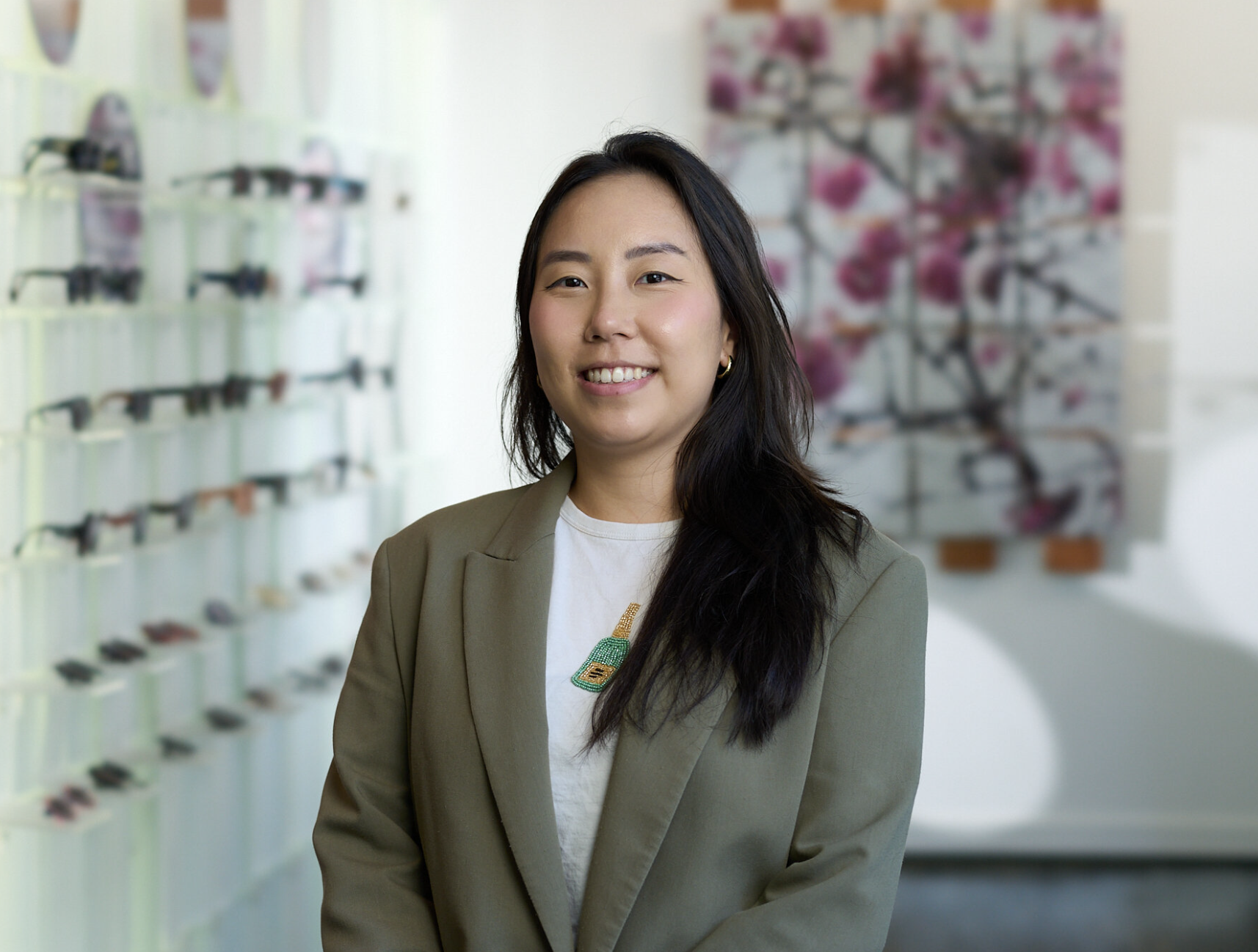 A woman with long dark hair, wearing a beige blazer and a t-shirt with a green graphic, standing inside a shop with eyewear displayed on the wall and a blurred background with pink flowers.
