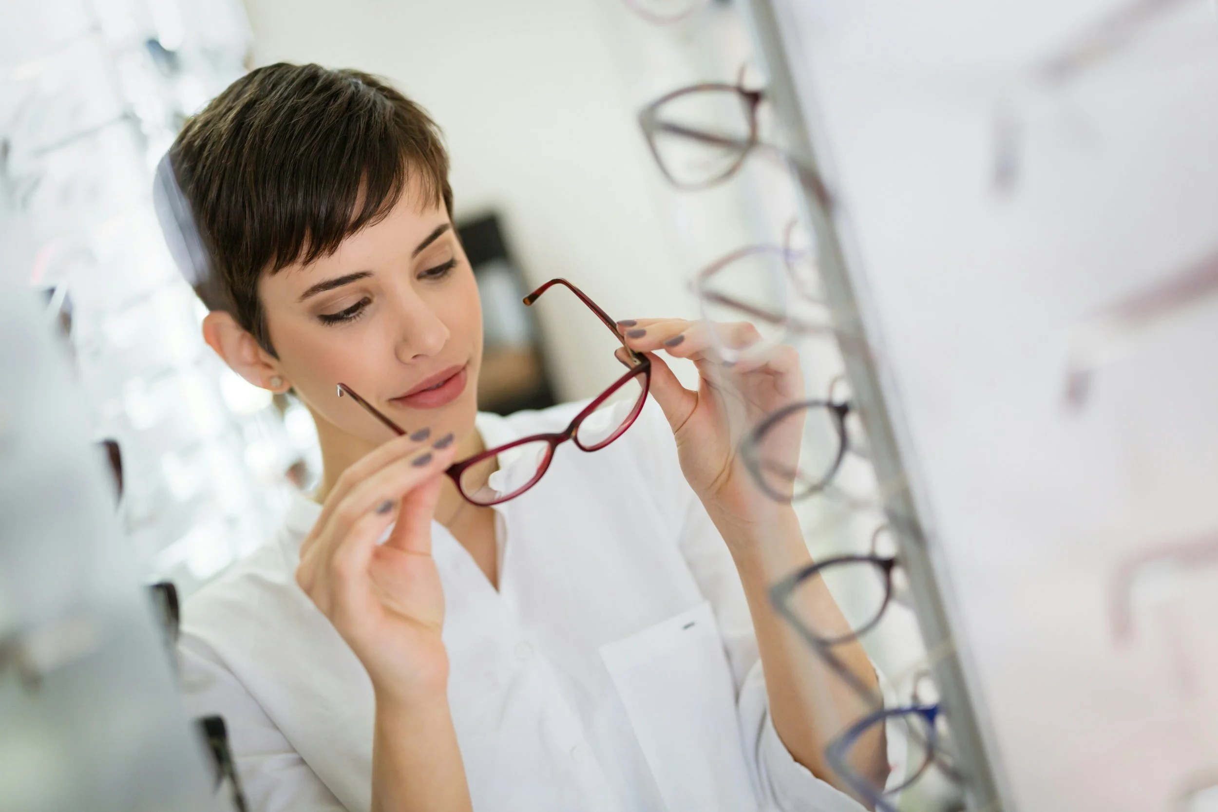 A woman with short dark hair and earrings is examining glasses while shopping for glasses in an optical store. She is holding a red-framed pair of glasses near her face, and there are multiple pairs of glasses on display on display racks.