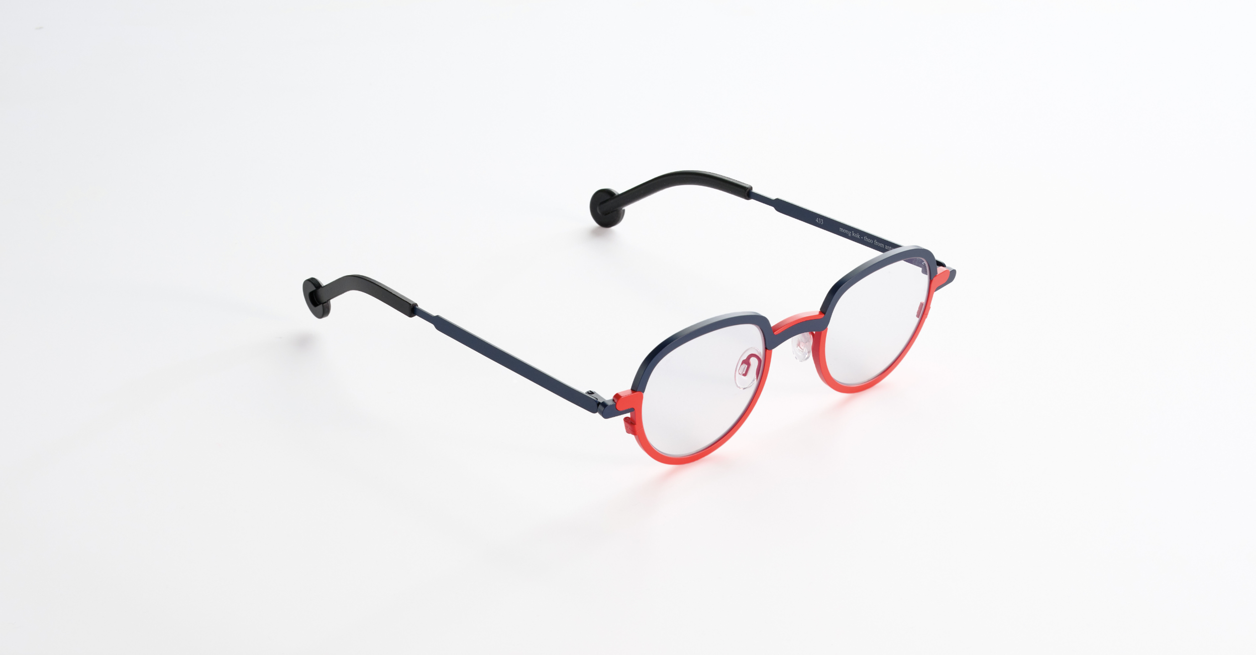 A pair of colorful Theo eyeglasses with a red frame, black temples, and clear lenses, resting on a white surface.