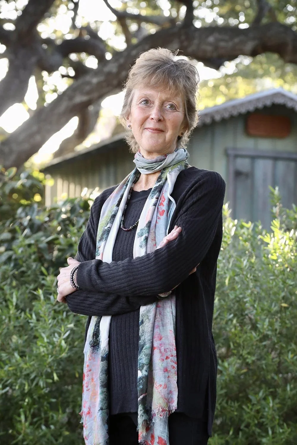 Amy Bacheller in her garden with greenery and a small green building, with sunlight filtering through the trees.