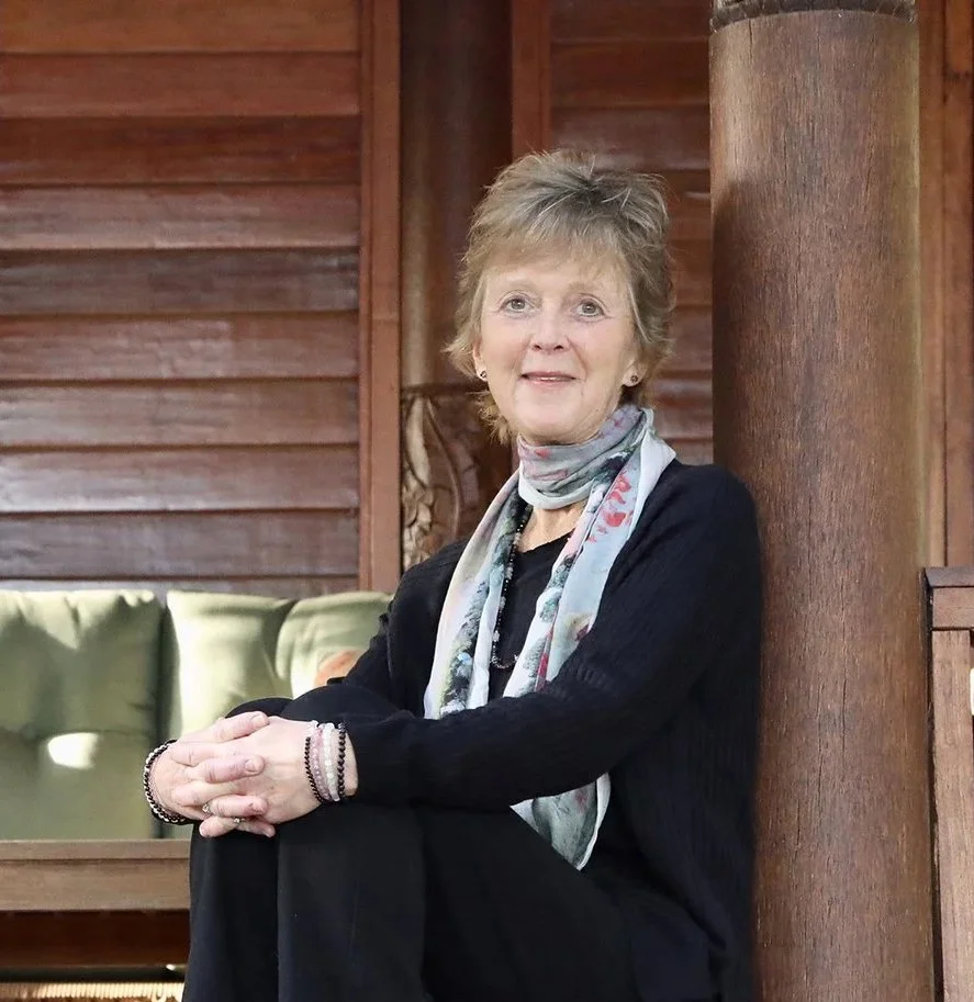Amy Bacheller in a colorful scarf around her neck, sitting on a wooden porch with wooden panel walls in the background.