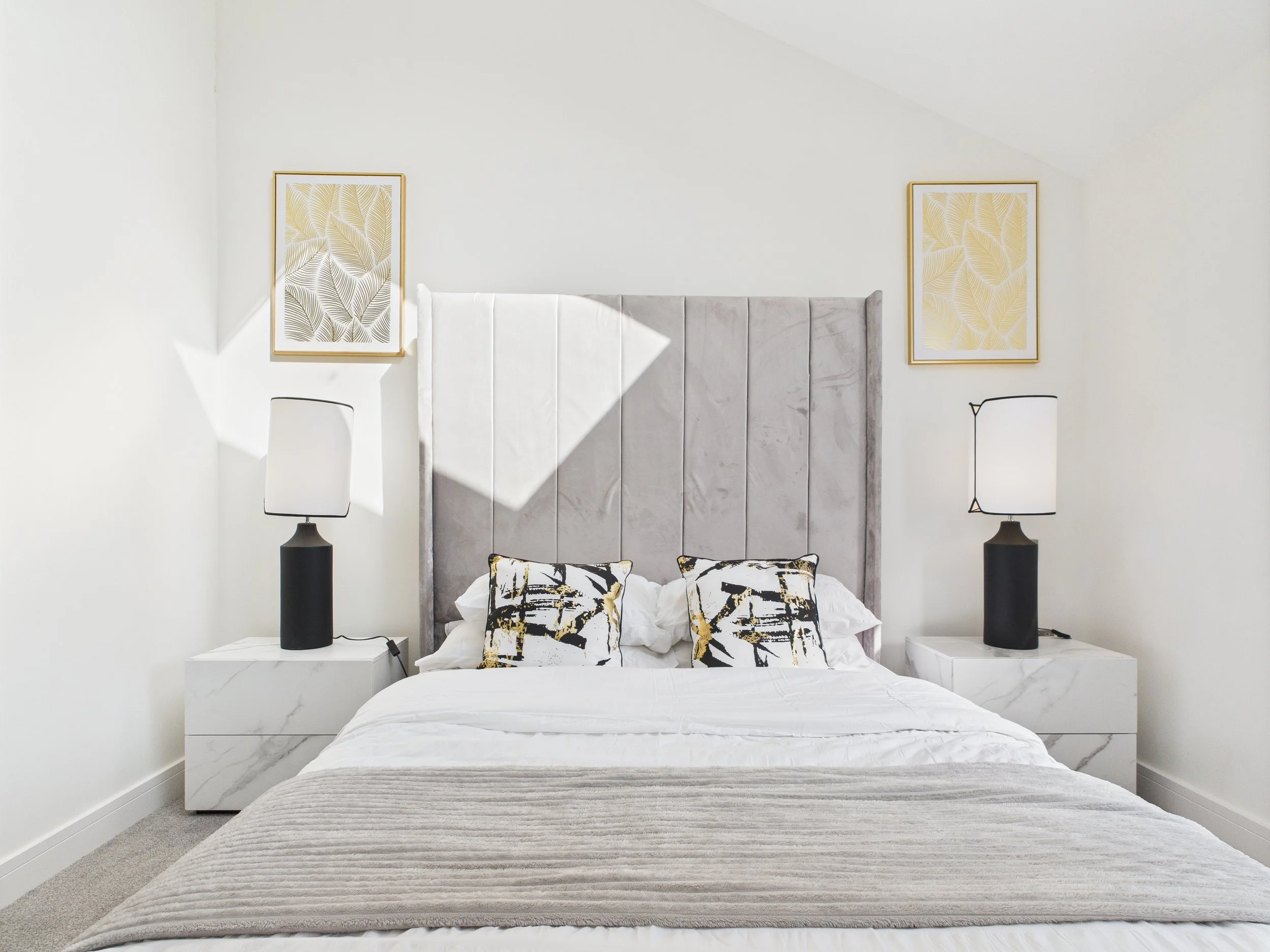 A modern bedroom with a large bed, gray velvet headboard, white bedding, black and yellow decorative pillows, two white marble nightstands, black and white table lamps, and framed leaf artwork on the walls.