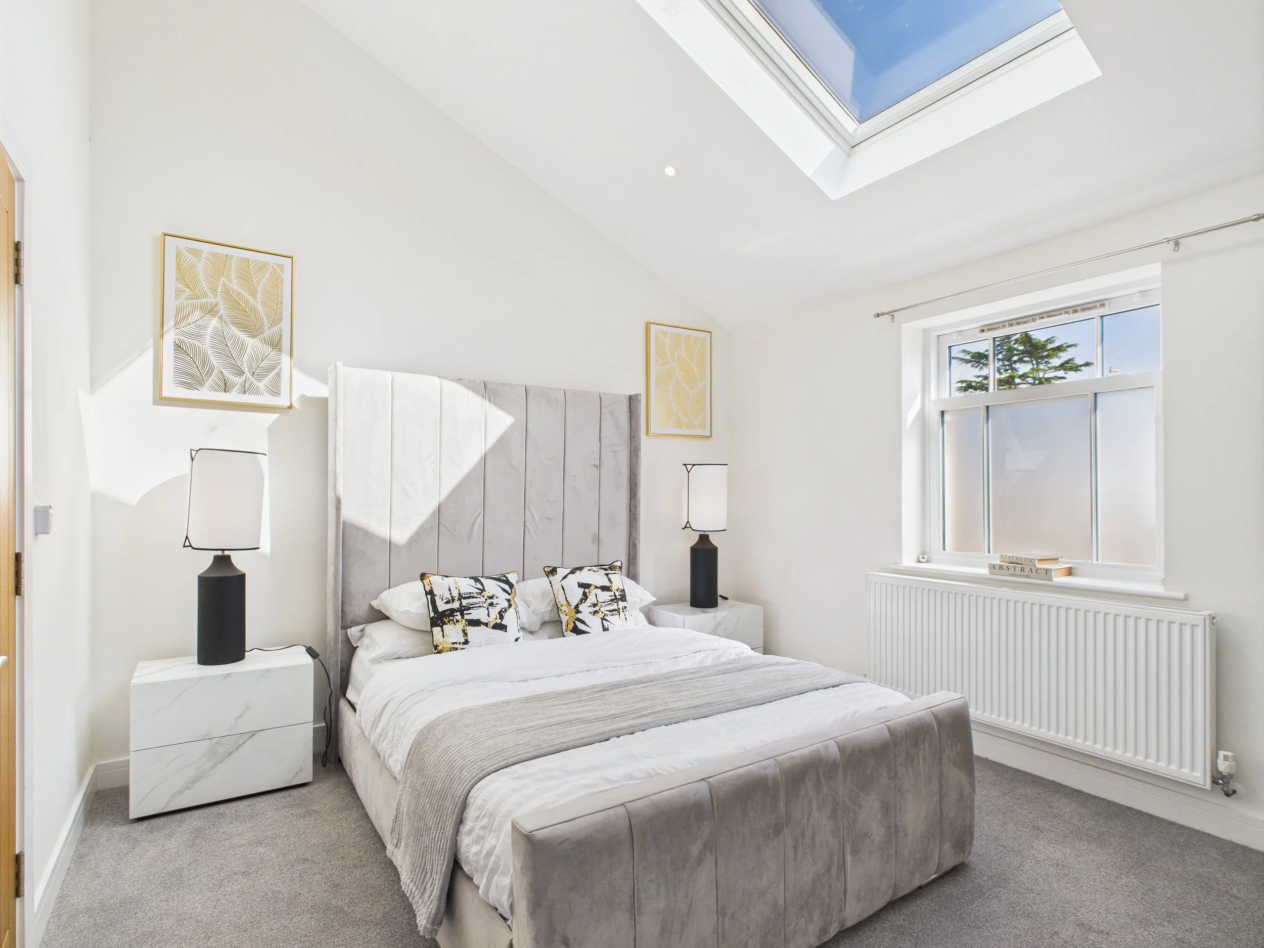 A modern bedroom with white walls, a large bed with a light gray upholstered headboard, white bedding, and black and white decorative pillows. There are two bedside tables with black lamps, artwork featuring yellow and green leaves on the walls, a window with a radiator below, and a skylight in the ceiling.