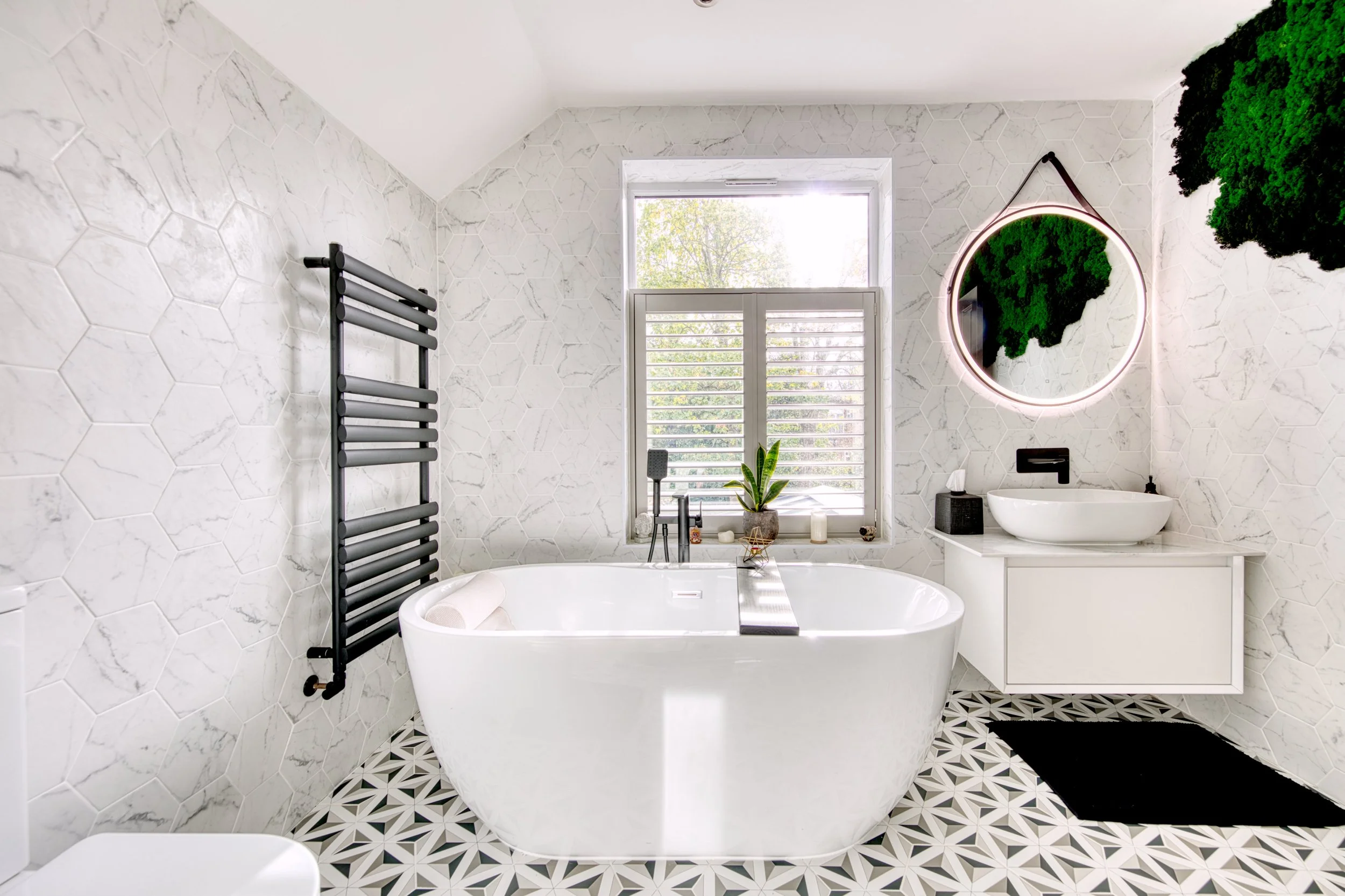 Modern bathroom with white freestanding bathtub, black heated towel rack, round mirror with pink LED light, vessel sink, and black toiletries, featuring geometric patterned floor and a window with white shutters.