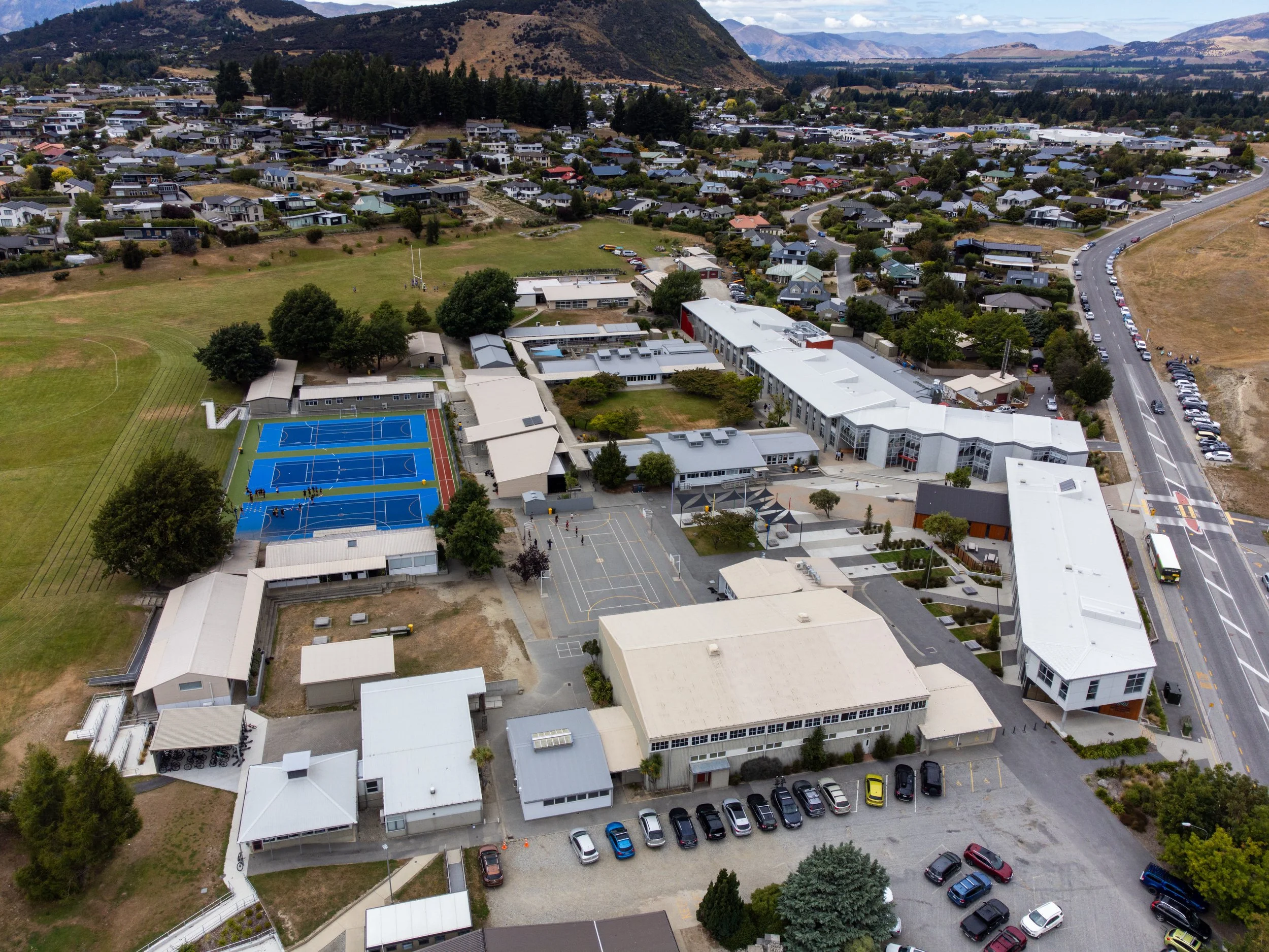 Aerial view of a school campus showing multiple buildings, tennis courts, a parking lot, a sports field, and residential neighborhood with mountains in the background.