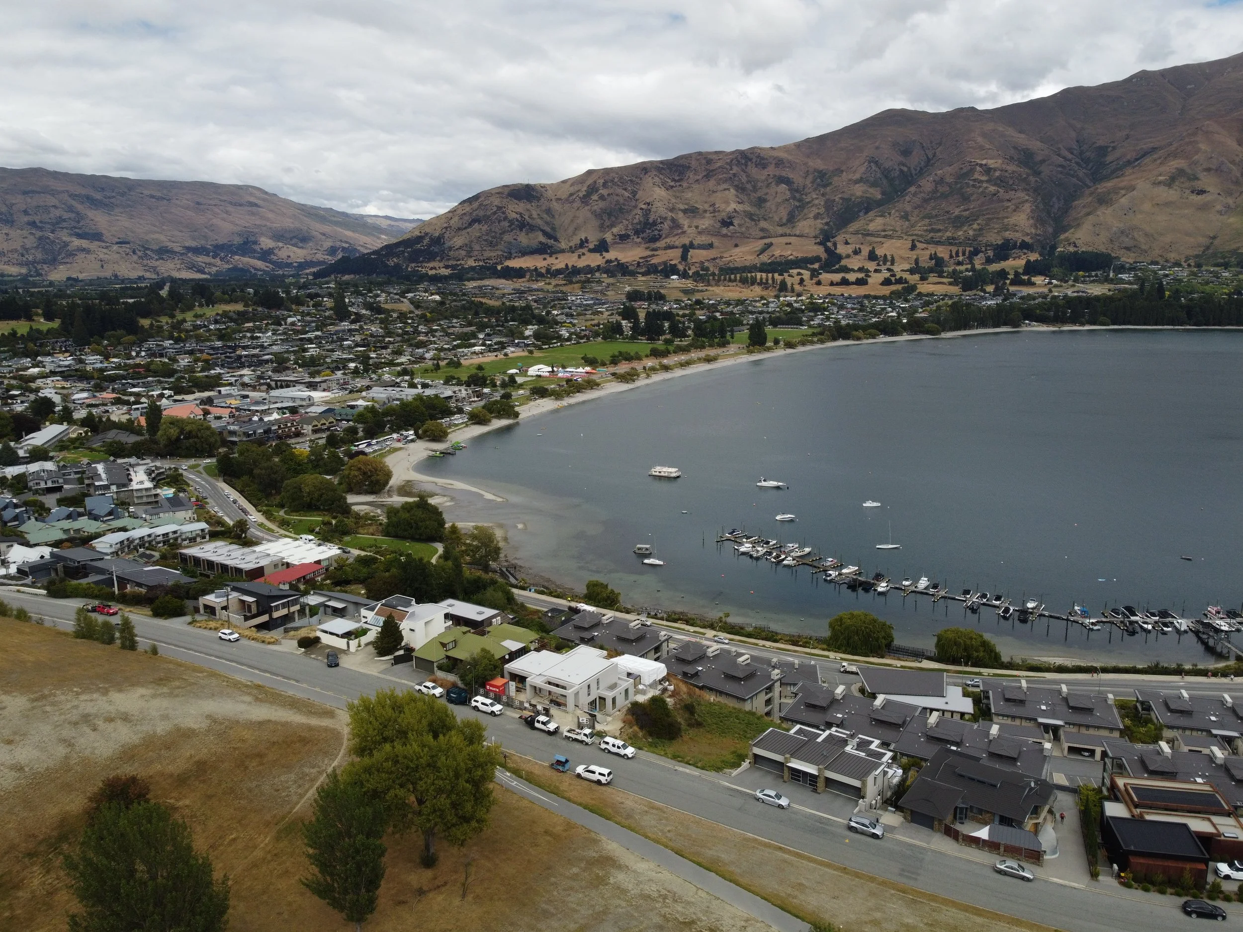 Aerial view of a lakeside town with boats docked at a marina, surrounded by residential houses and hills in the background.