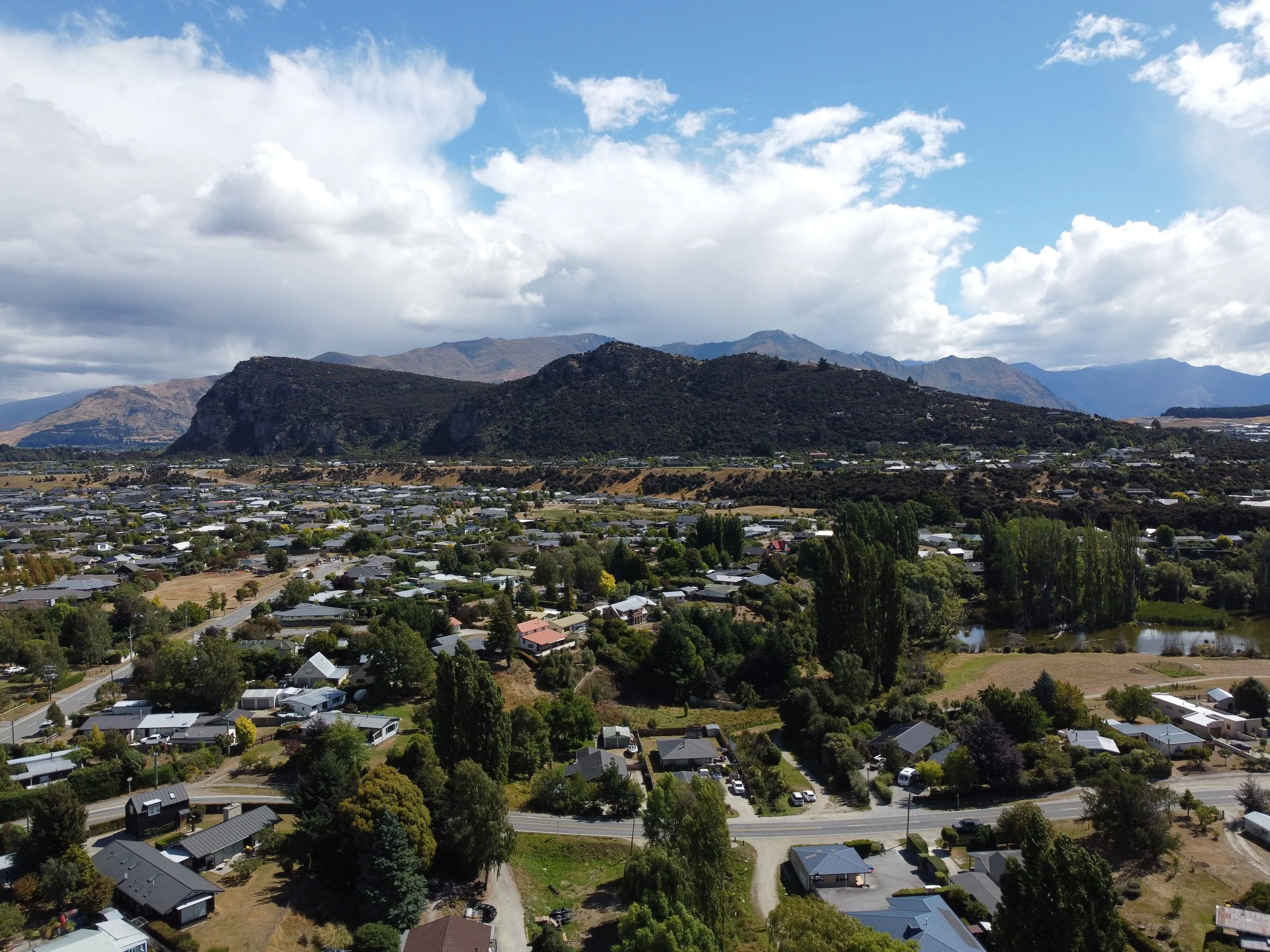 Sky shot,  of a small town with many houses, trees, and a small pond, set against a backdrop of mountains under partly cloudy skies.