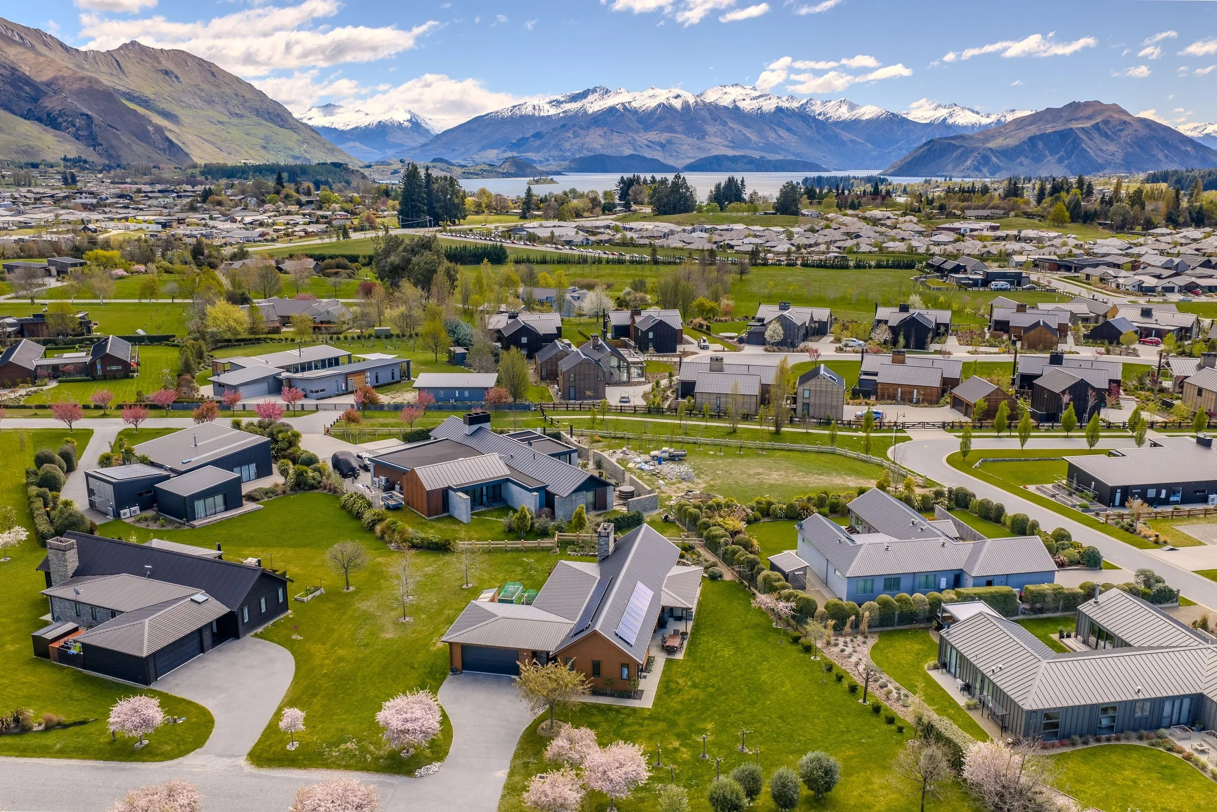 Aerial view of a residential neighborhood with modern houses, lush green lawns, and blooming trees, set against snow-capped mountains and a lake in the background.