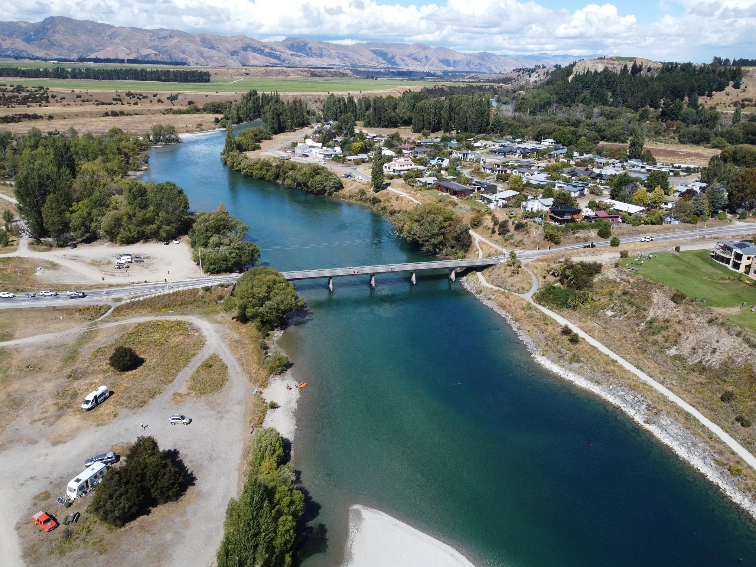 sky shot , of a river flowing through a town, with a bridge crossing over it, surrounded by houses, trees, and mountains in the background.