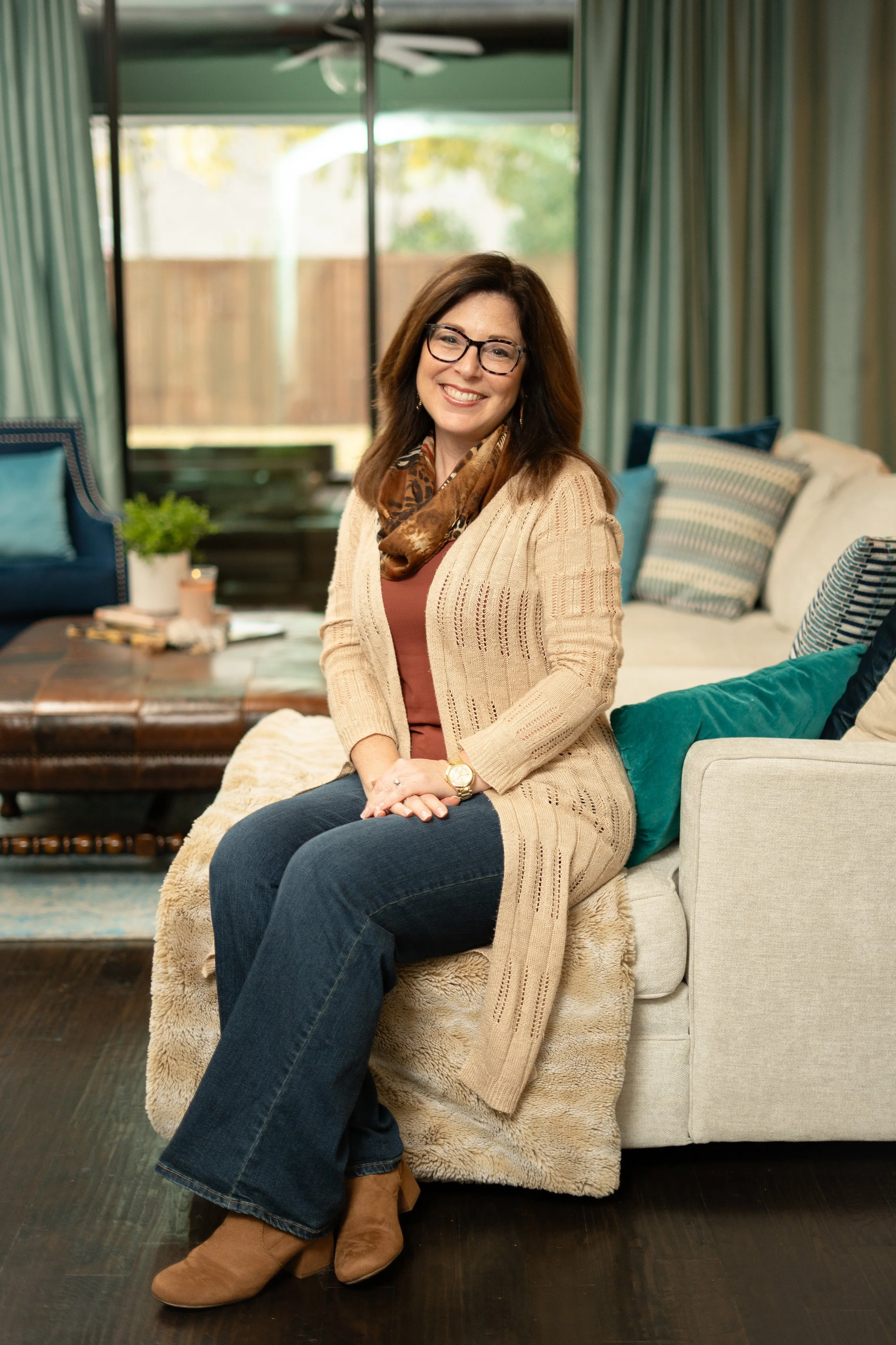 A woman with glasses, brown hair, and a warm smile, sitting on a sofa in a cozy living room with colorful pillows and a large window in the background.
