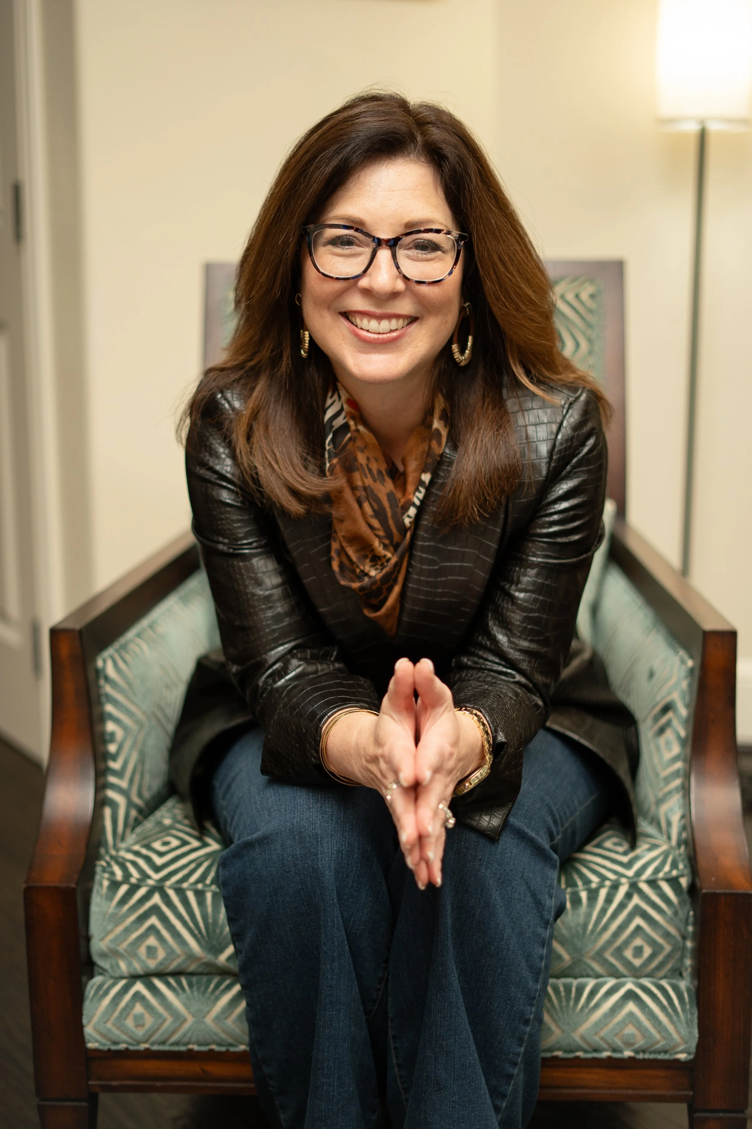 A woman with brown hair, glasses, and a bright smile sitting on a patterned chair, wearing a leather jacket, scarf, and jewelry, in a warmly lit room.