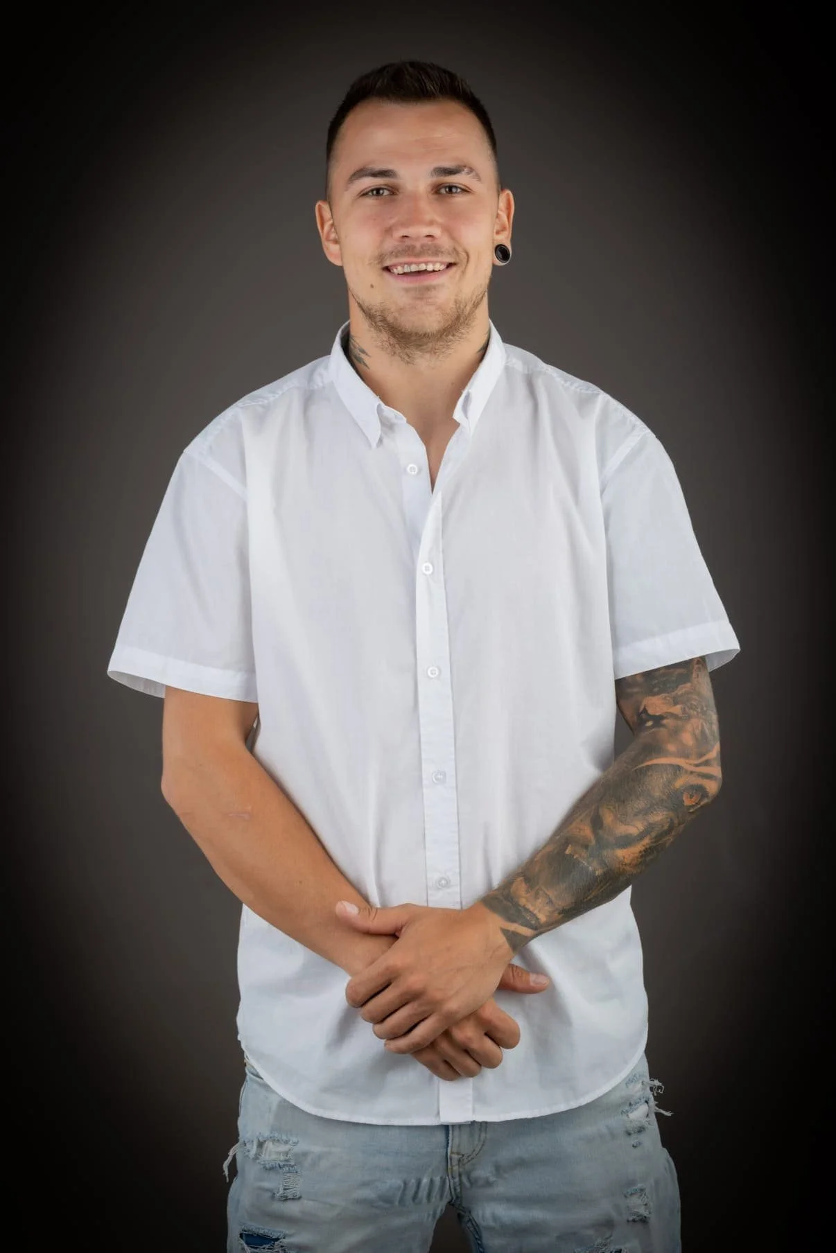 Junger Mann, weißen Hemd und Jeans vor dunklem Hintergrund, lächelnd.