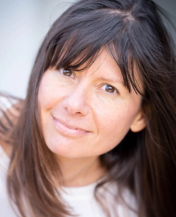 Close-up of a smiling woman with dark brown hair and light skin, wearing a white top.