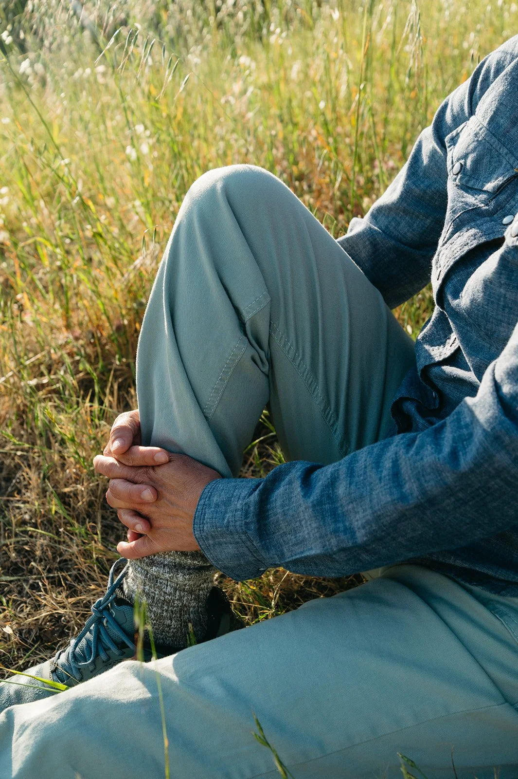 Person sitting in a grassy field holding their knee, wearing a denim jacket, light-colored pants, and hiking shoes.