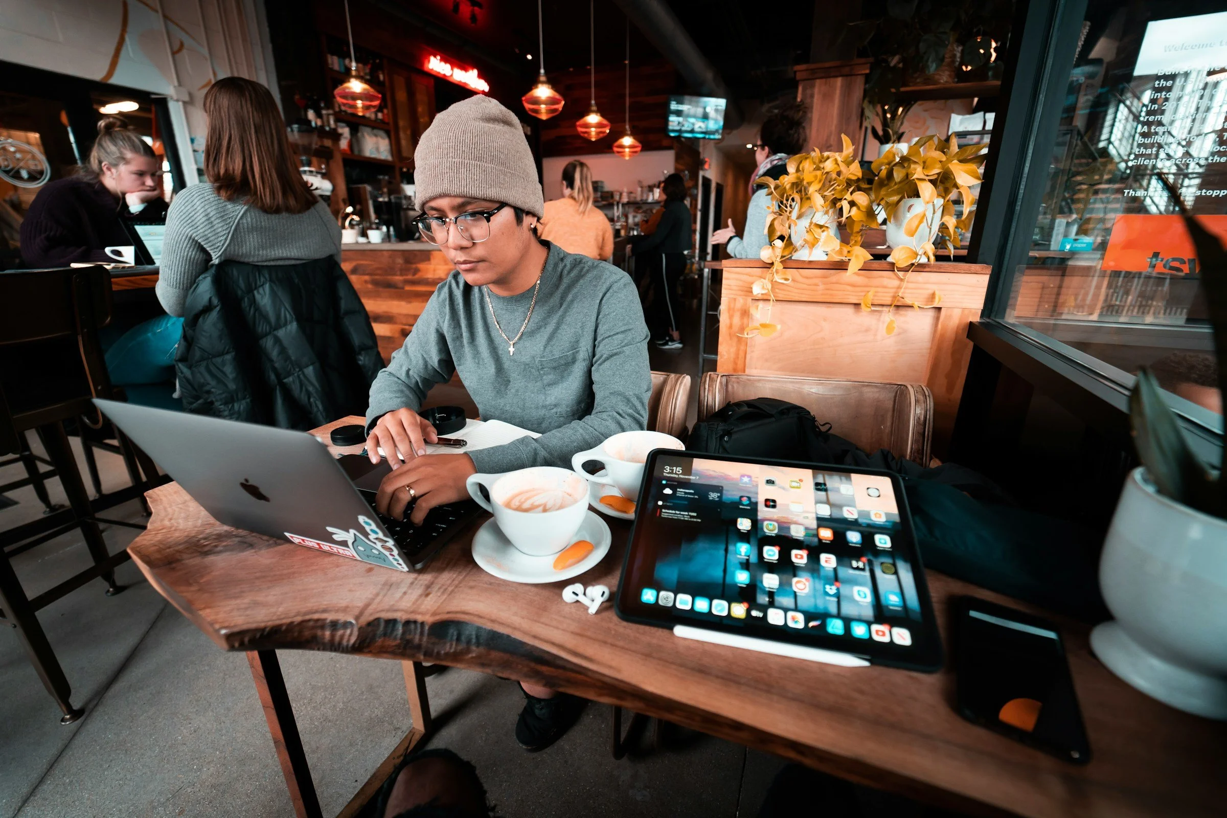 a young man works on his computer in a bustling coffee shop, with multiple tech devices on his table