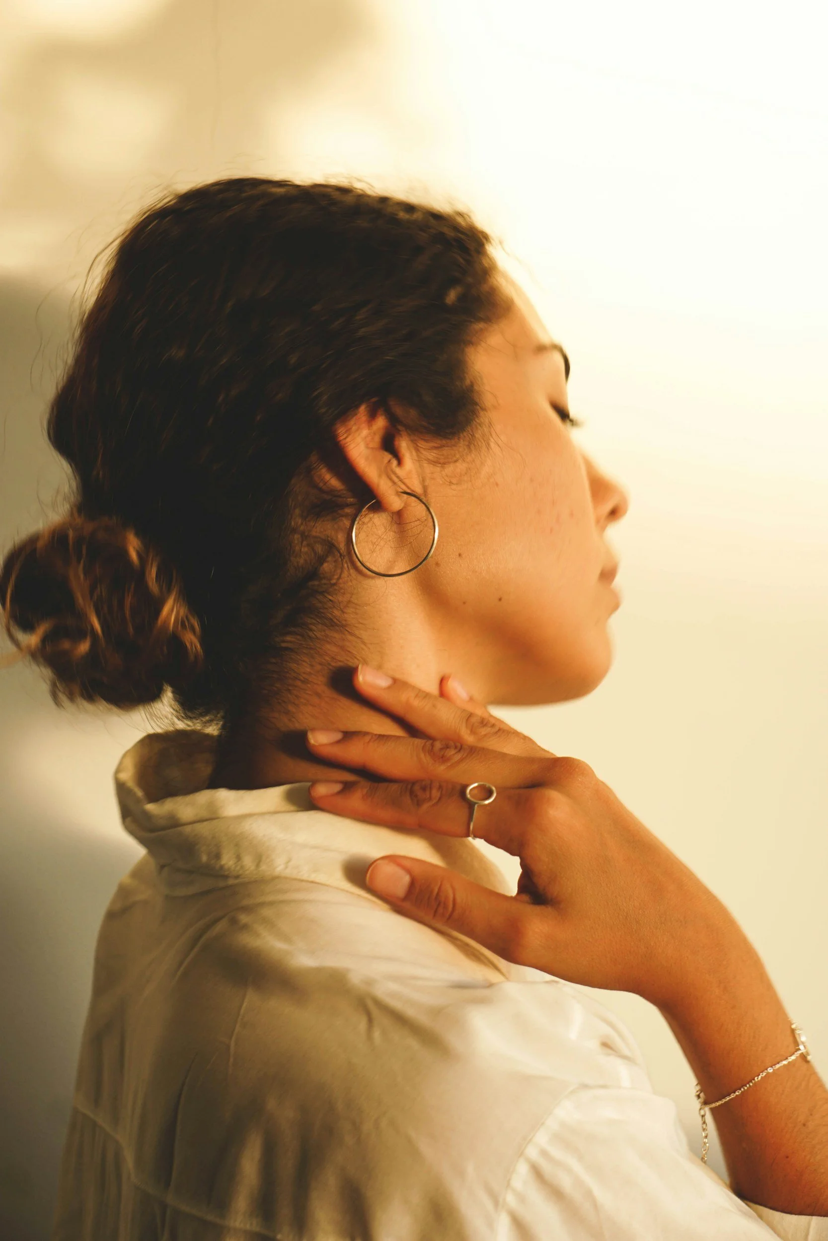 A woman with dark hair tied in a bun, wearing hoop earrings, rings, and bracelets, is seen in profile, with her hand gently touching her neck against a warm, softly lit background.