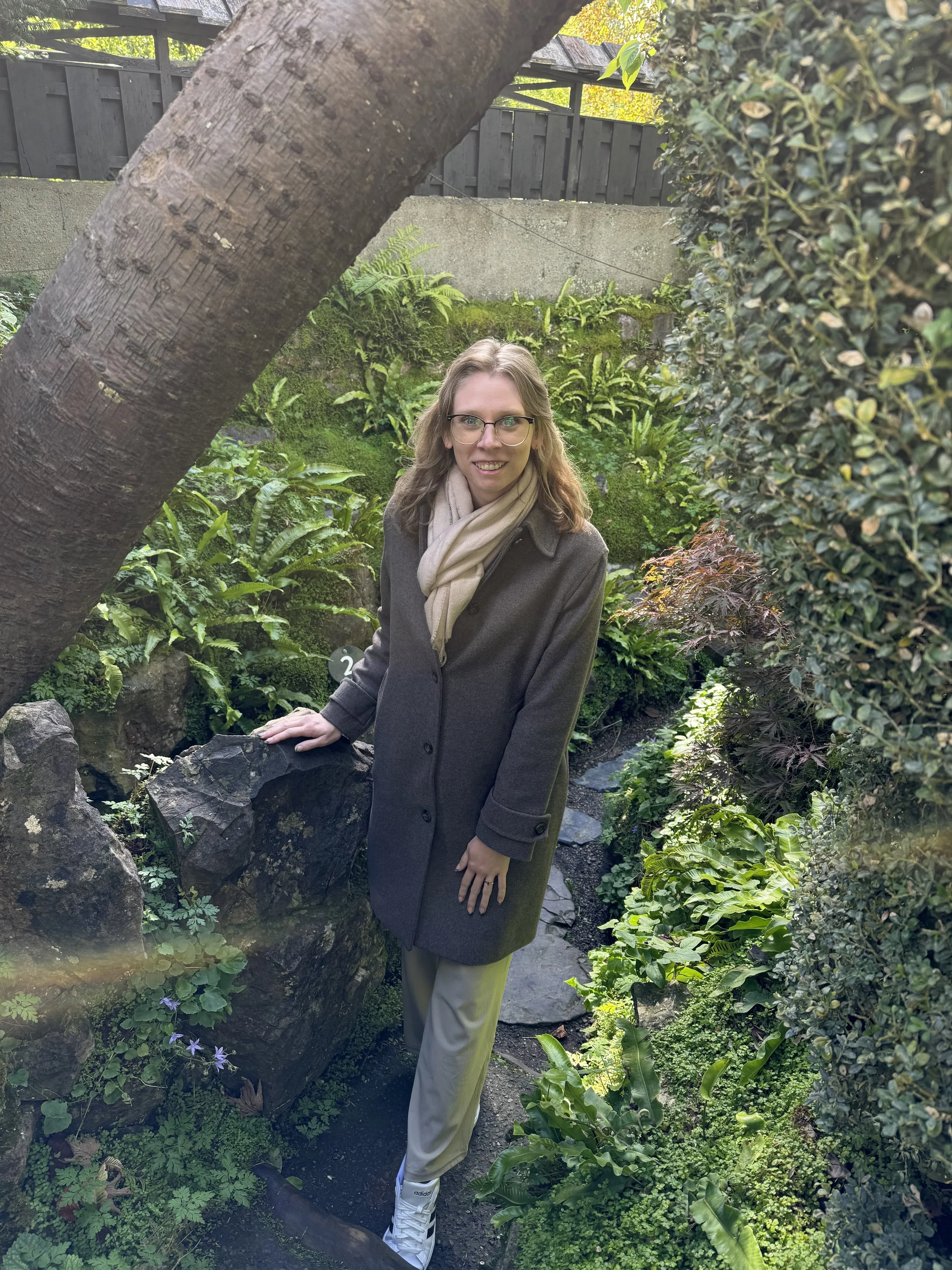 A woman standing amidst lush greenery in a garden, leaning on a rock, wearing glasses, a dark coat, beige pants, a light-colored scarf, and white sneakers.
